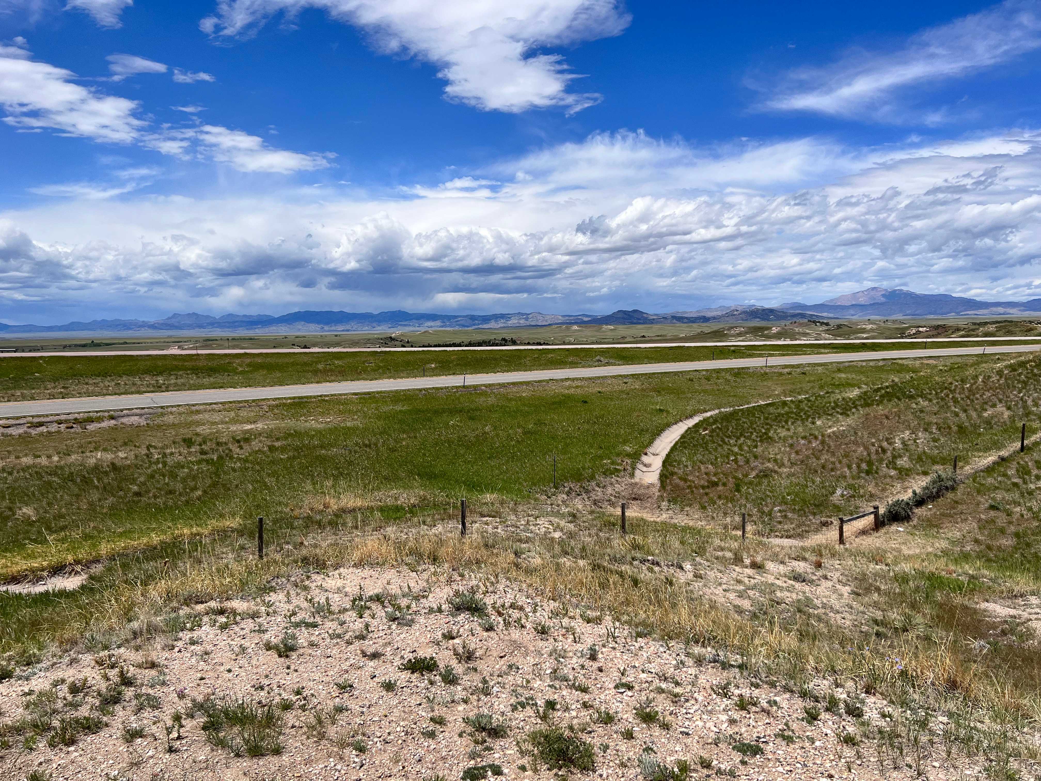 A highway running across a grassy plain, with the large, rocky Laramie Mountains in the background, and the shadows of a sky full of fluffy white clouds reflecting on their peaks.