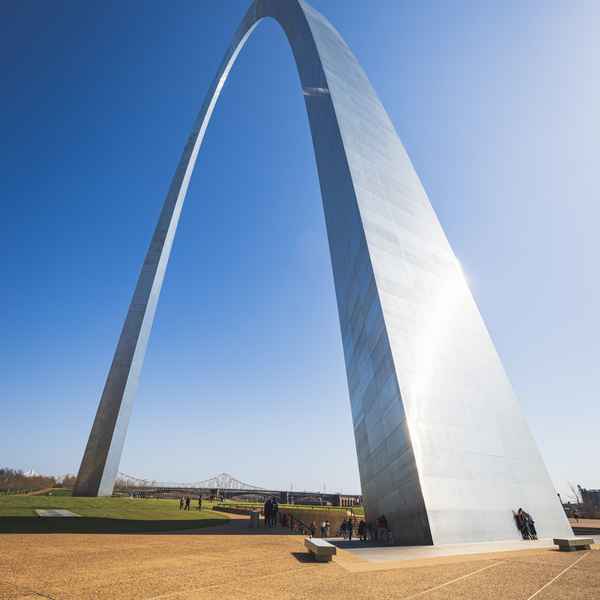 Two people lean against the side of the massive, silvery metal Gateway Arch as it towers into the blue sky
