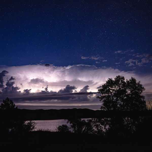 Fluffy clouds are lit by lightning flashes in front of a deep blue sky filled with stars