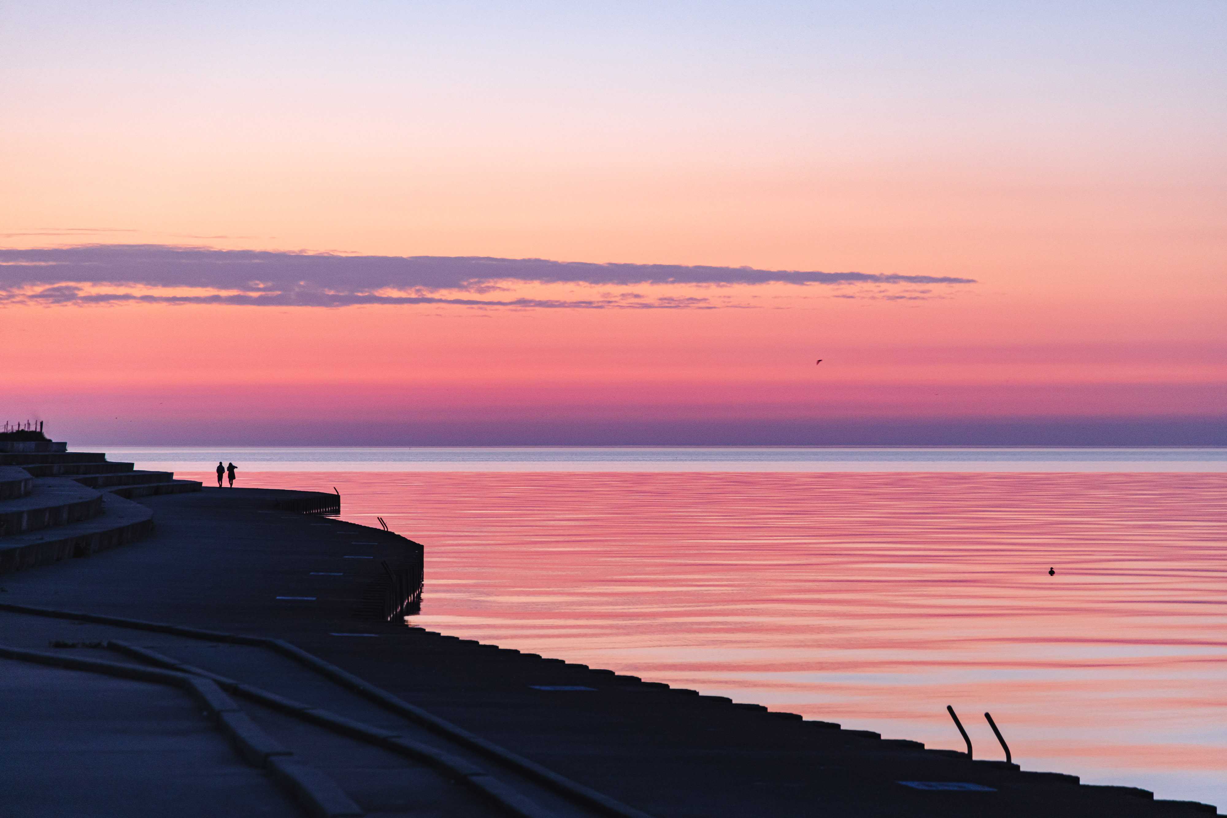 The sky is filled with orange and pink tones, blending smoothly into the gentle waves of Lake Michigan where water meets sky. To the left of the image, the silouhette of a couple walking into the sunrise along the sharp lines of the concrete revetments that line the lakeshore. 