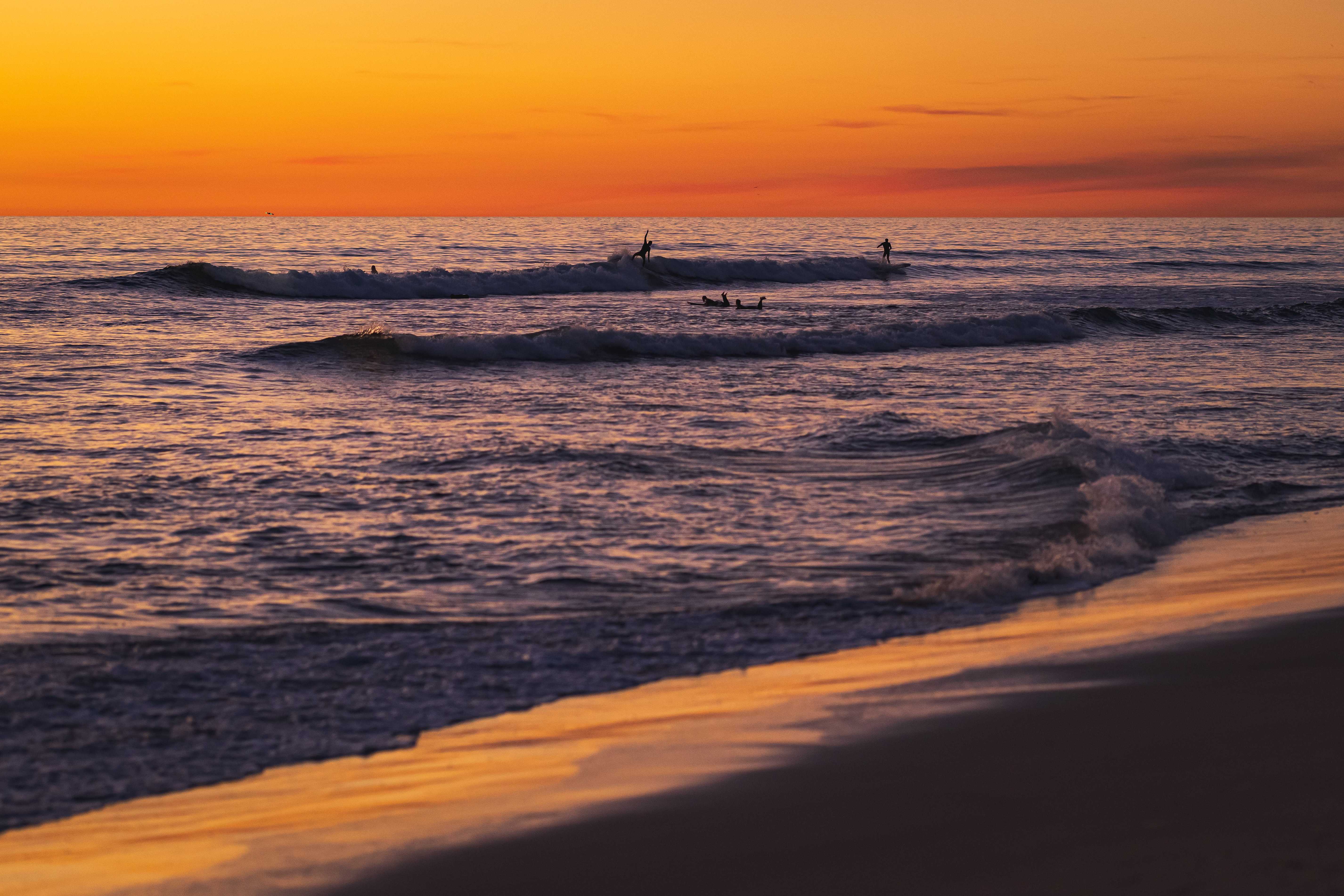 A deep orange and yellow sunset highlights a deep blue sea. Surfers are riding the last waves of the day.