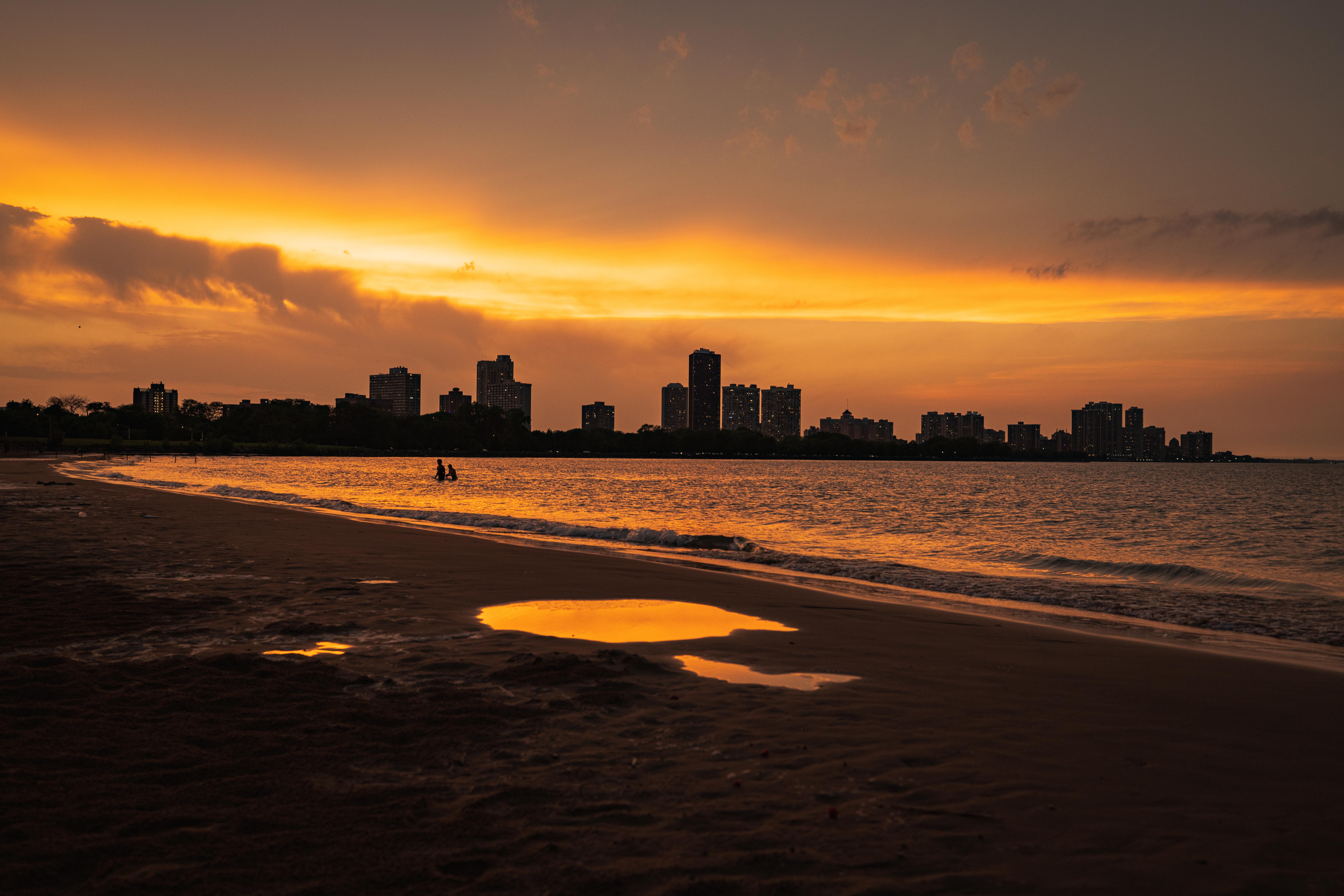 Two people walk toward shore from shallow water while a dramatic orange sunset reflects on the water
