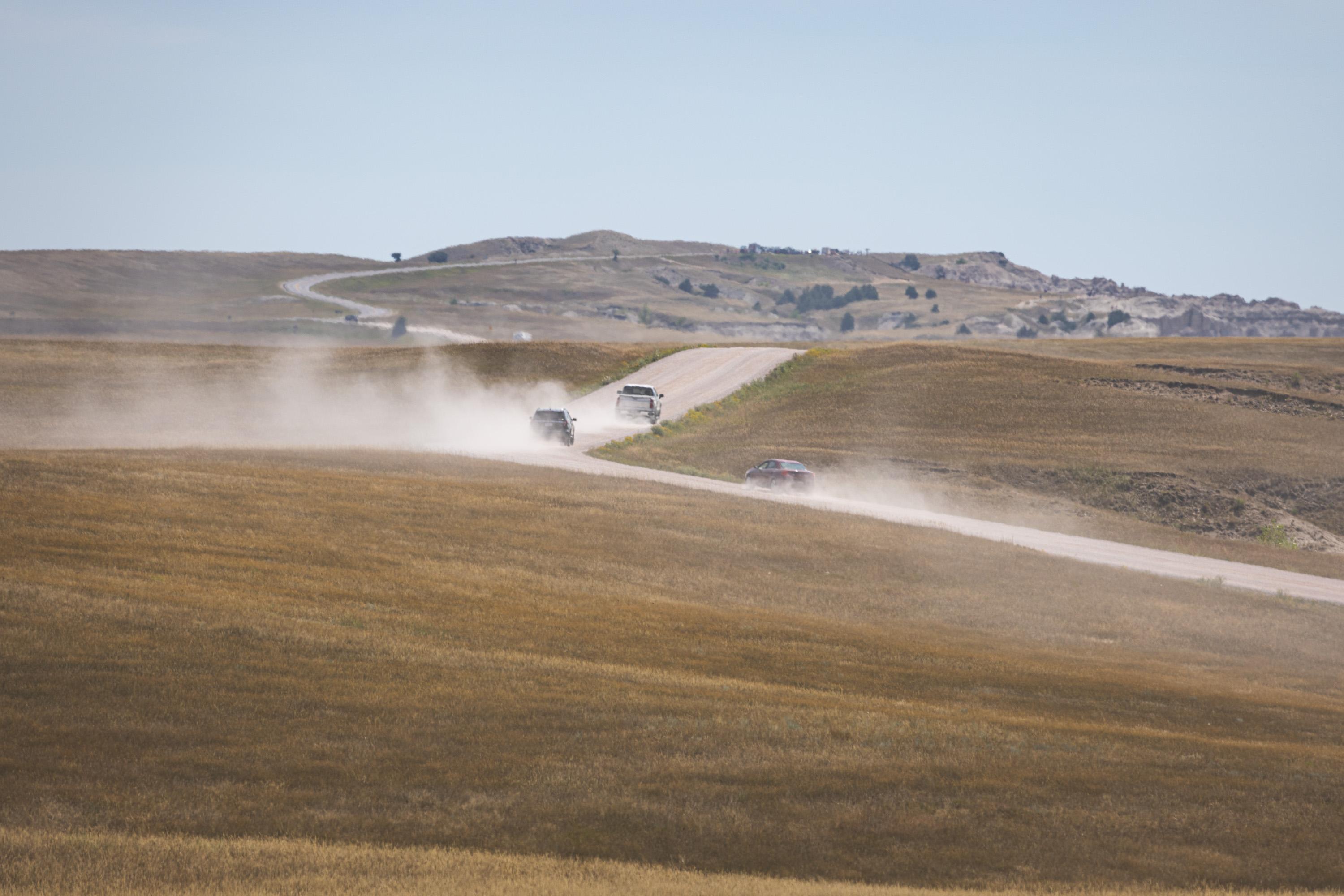 Cars in the park wind up a crushed gravel dirt road as they kick up dust clouds behind them