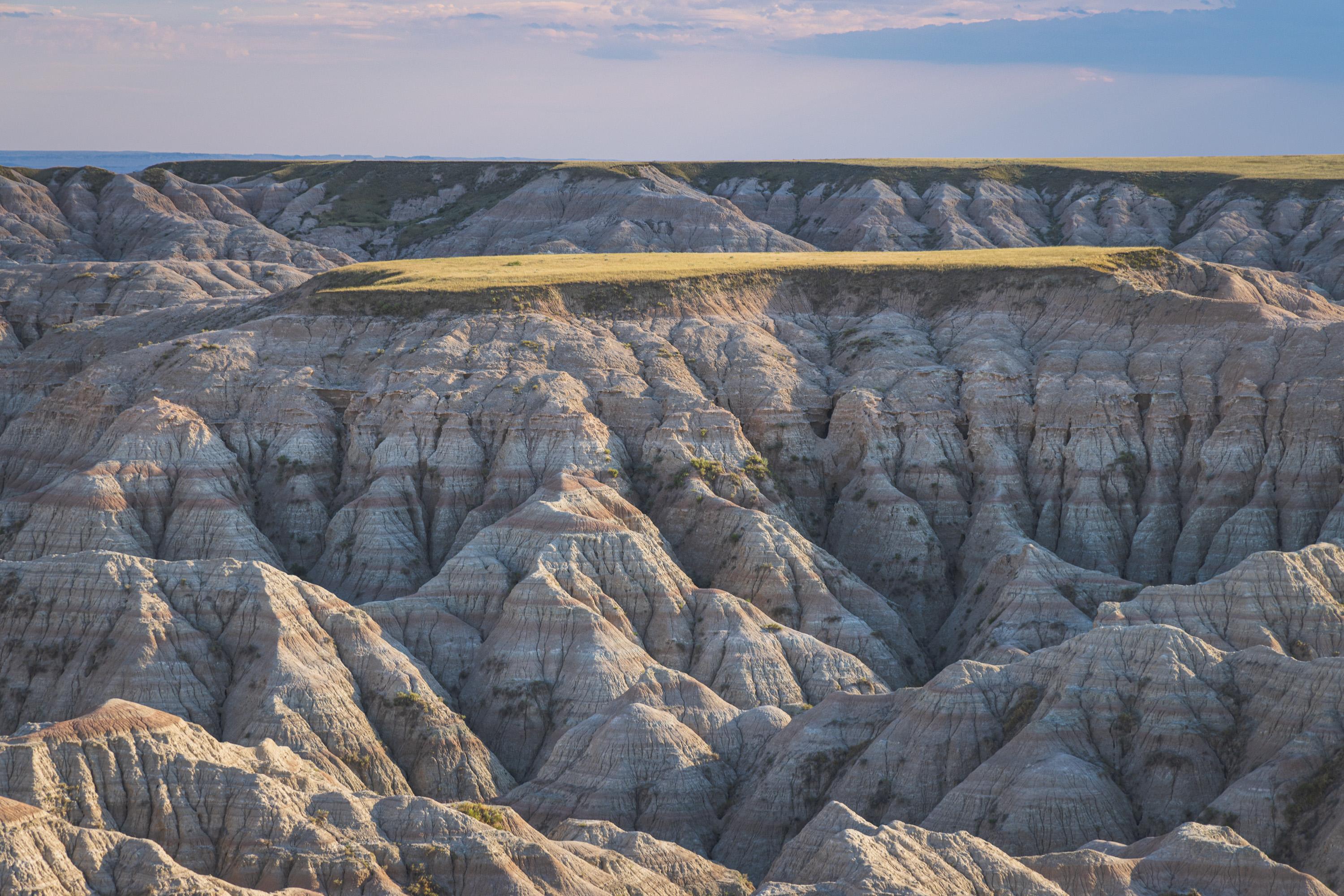 A plateau of soft green grass above a valley showing dramatic, layered rocks in the soft evening light