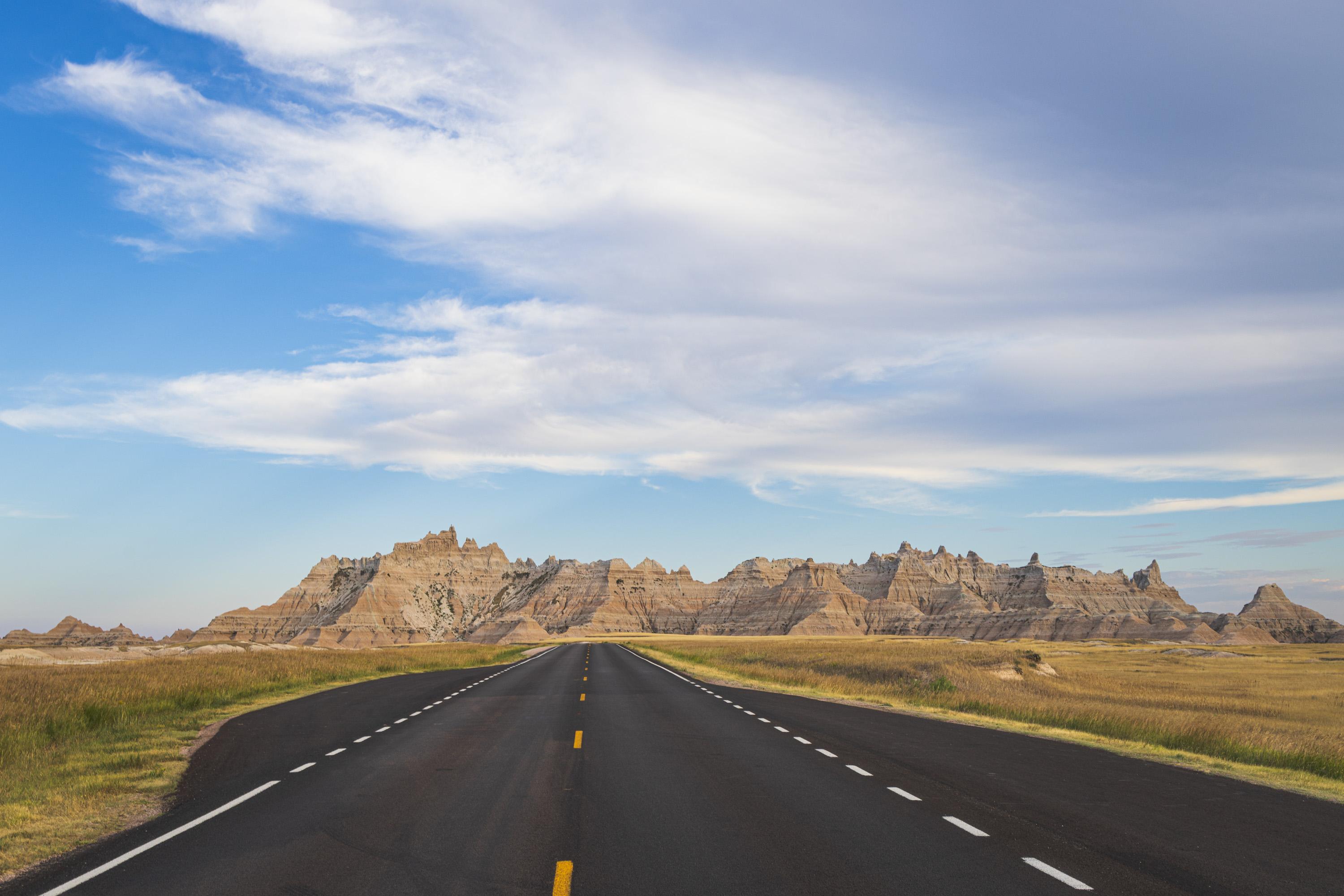 A dramatic vista as a road stretches toward a sharp mountain of rock as dark cloud cover blows in from the right of the image. The road is the same one shown in the previous photo.