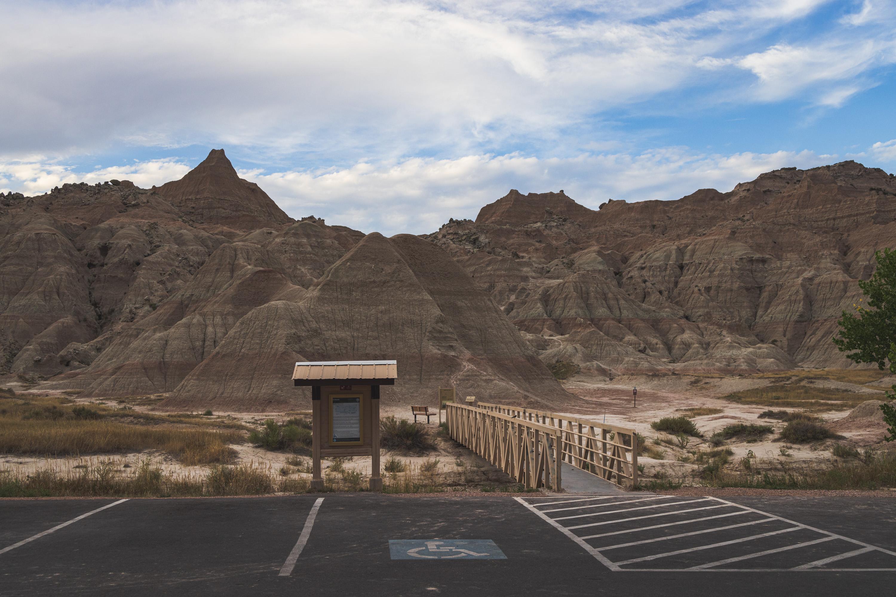 Standing in a small parking lot looking at the start of a trail, which crosses a small bridge next to a signpost and leads into the rocky hills which I had just been climbing