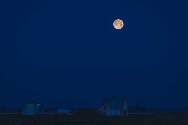 The bright, full moon provides a dull shine on a deep blue night revealing farm buildings in a field