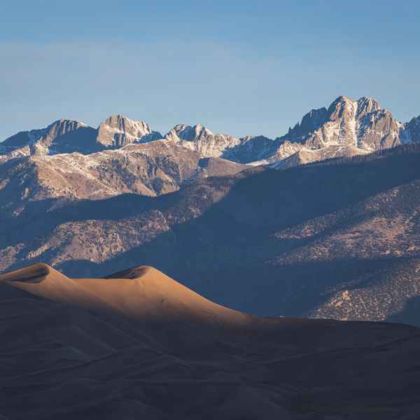 A massive, rolling sand dune is lit just on its highest curves as the sun rises, and behind it, a line of high, sharp, craggy mountaintops are being illuminatd, revealing snowcapped peaks