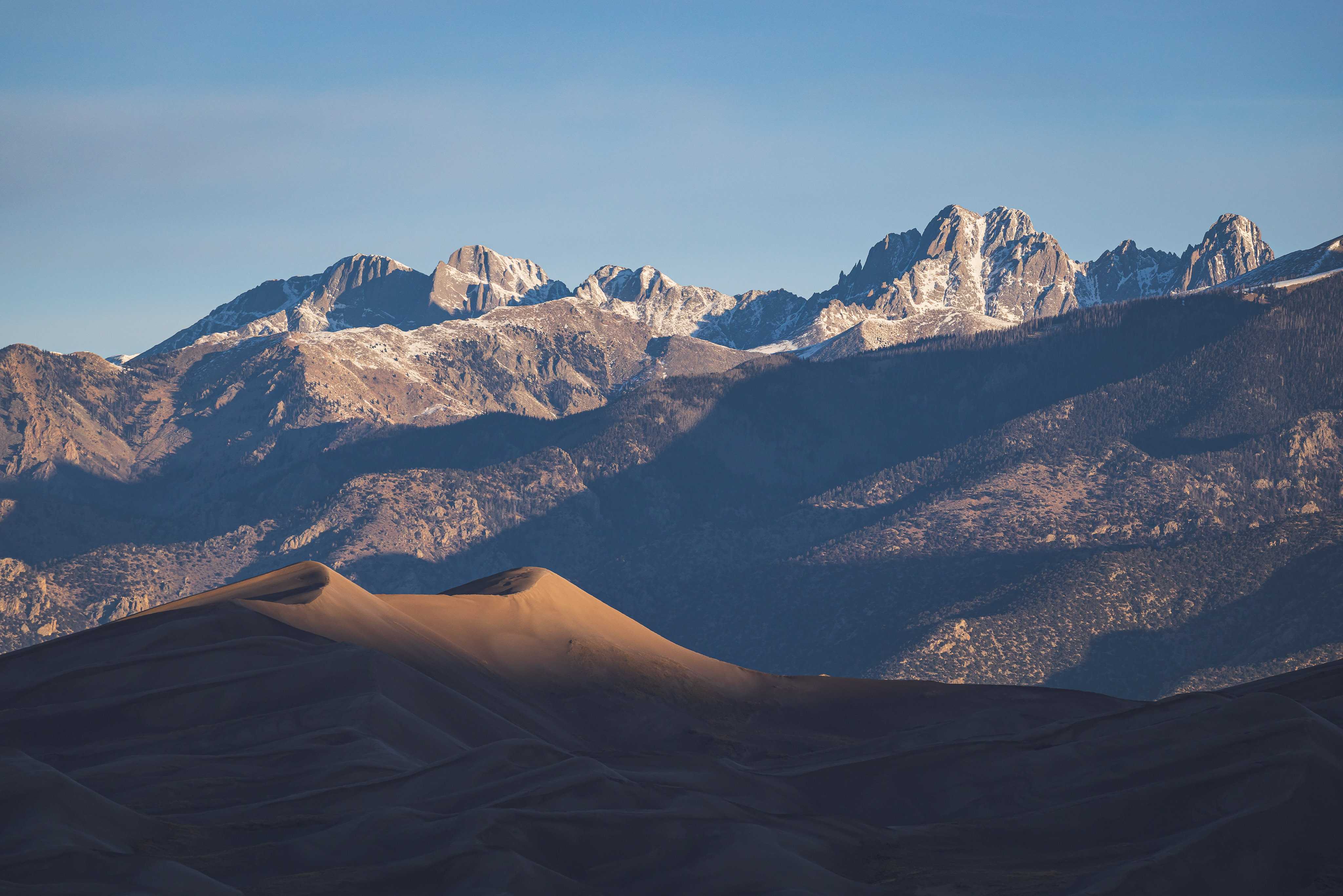 A massive, rolling sand dune is lit just on its highest curves as the sun rises, and behind it, a line of high, sharp, craggy mountaintops are being illuminatd, revealing snowcapped peaks