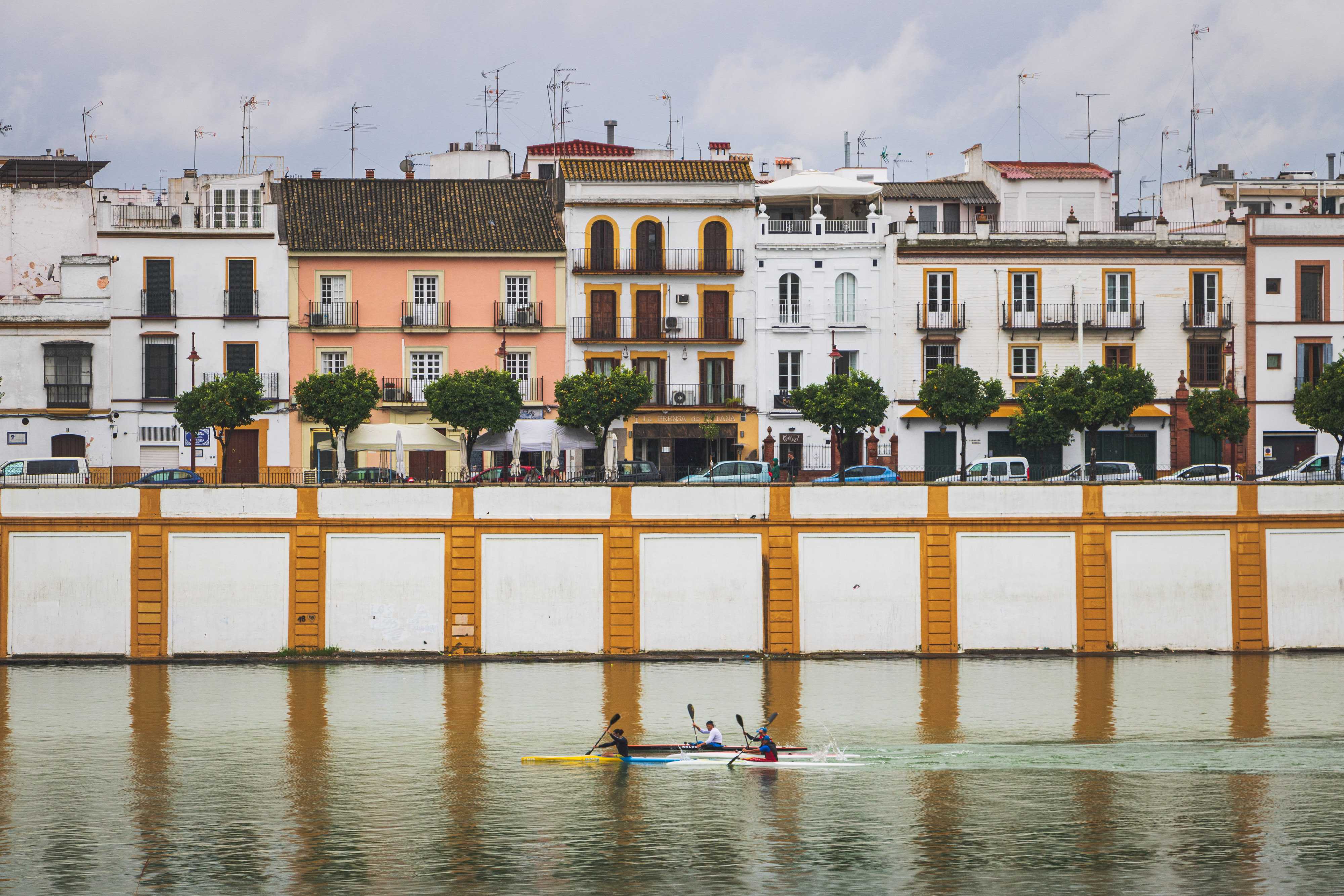 Three men on small kayaks are padding down a river, while behind them the white painted river wall and colorful tile-roofed buildings of Triana, one of the oldest neighborhoods in Sevilla, Spain.