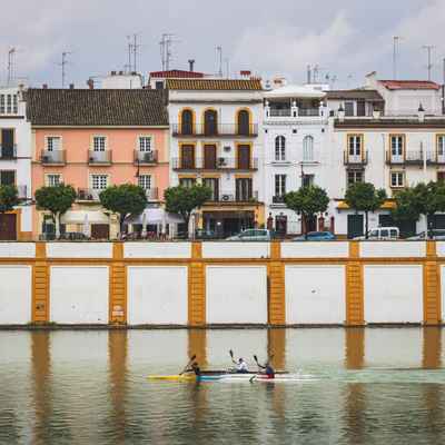 Three men on small kayaks are padding down a river, while behind them the white painted river wall and colorful tile-roofed buildings of Triana, one of the oldest neighborhoods in Sevilla, Spain.