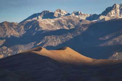 A massive, rolling sand dune is lit just on its highest curves as the sun rises, and behind it, a line of high, sharp, craggy mountaintops are being illuminatd, revealing snowcapped peaks