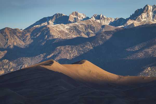 A massive, rolling sand dune is lit just on its highest curves as the sun rises, and behind it, a line of high, sharp, craggy mountaintops are being illuminatd, revealing snowcapped peaks