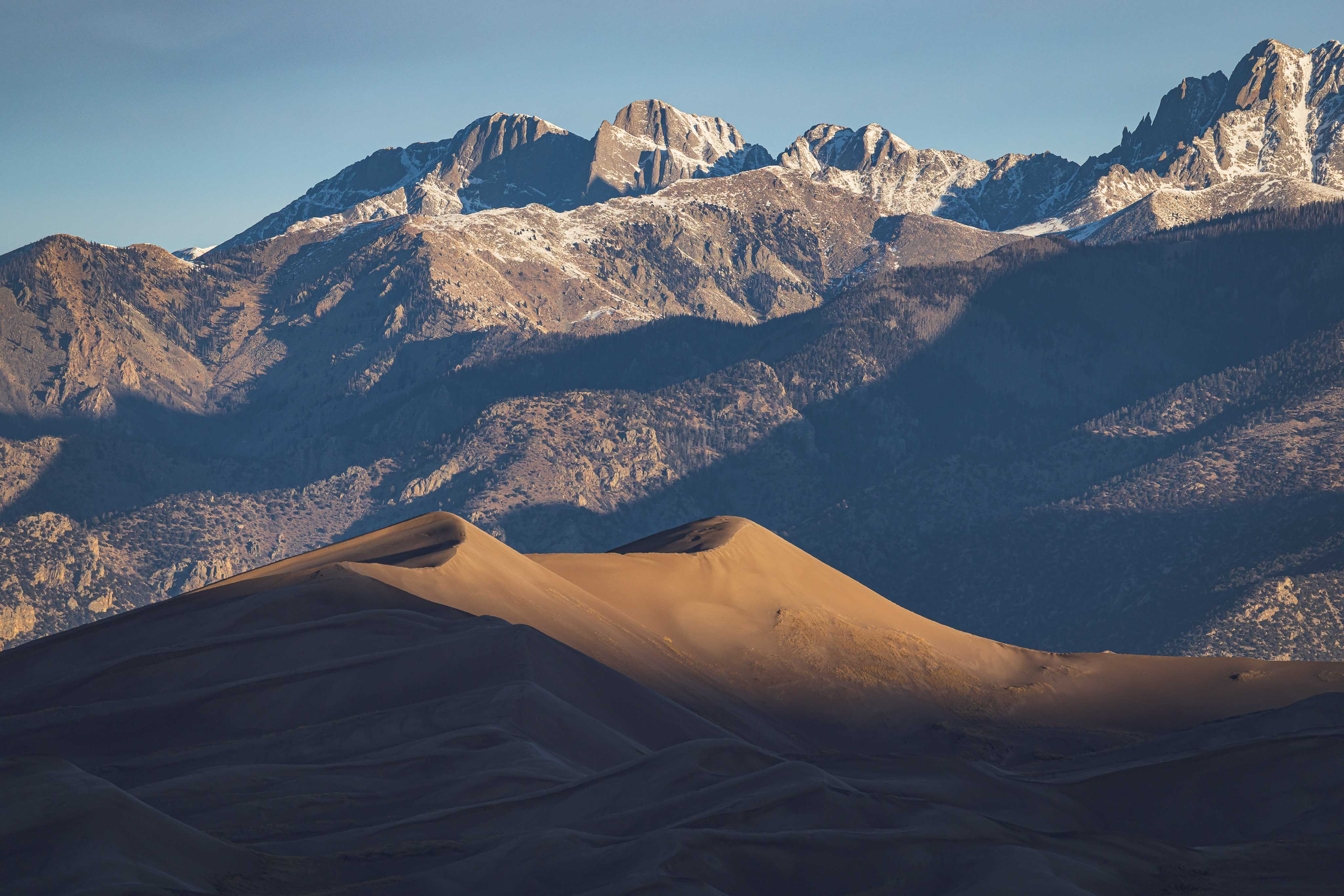 A massive, rolling sand dune is lit just on its highest curves as the sun rises, and behind it, a line of high, sharp, craggy mountaintops are being illuminatd, revealing snowcapped peaks