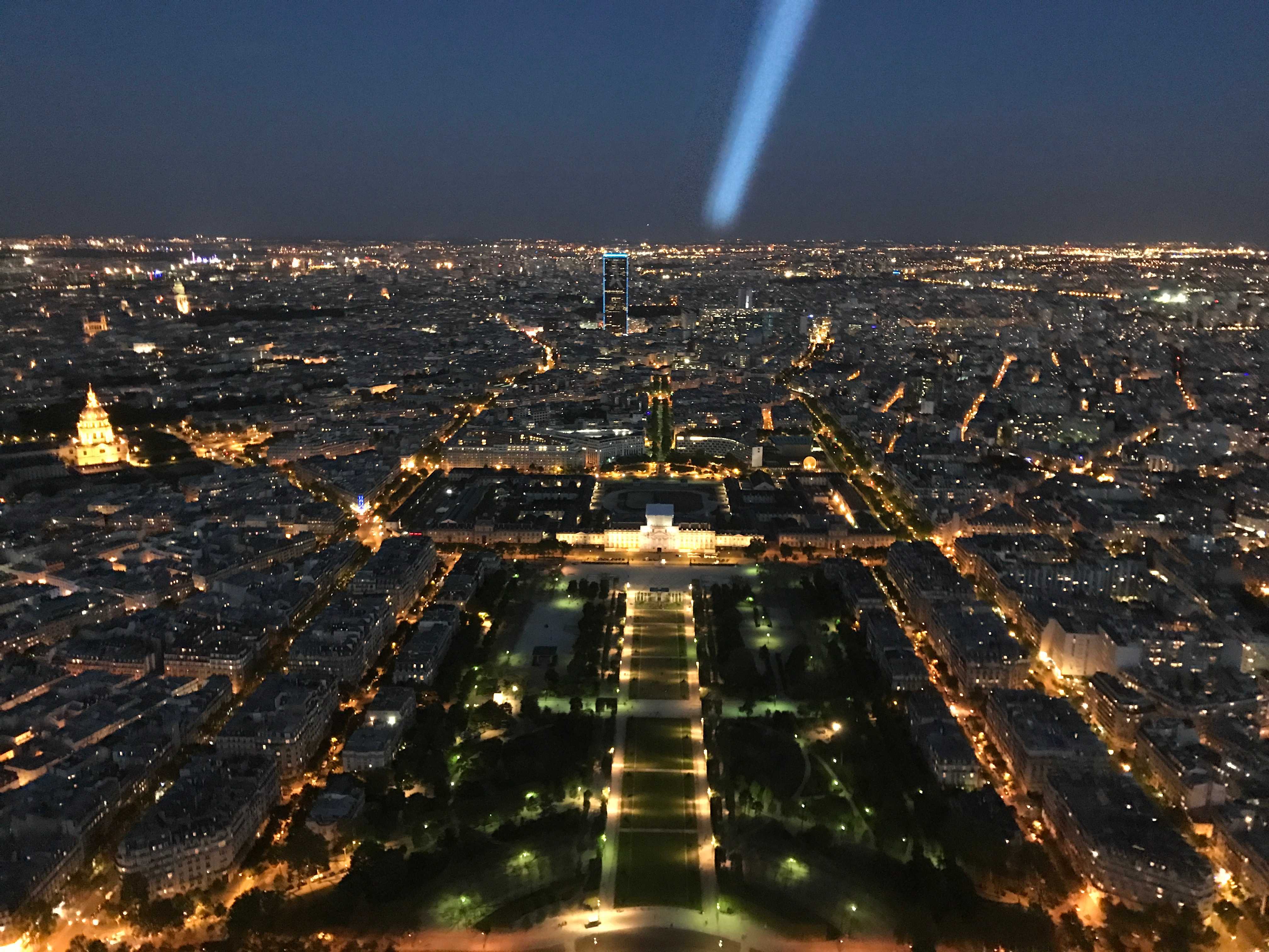 The city of Paris at night, viewed from above at the top of the Eiffel Tower; a dark green park is punctured by golden streetlights, surrounded by many city streets lit by buildings and streetlamps. A bright beam of light shines from overhead toward the horizon.