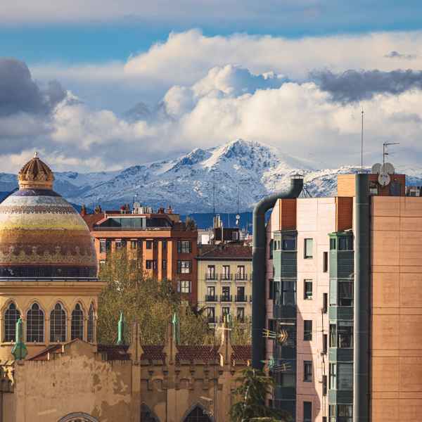 A snow-covered mountain shrounded in white fluffy clouds rises behind the earth-colored tile and paint of Madrid rooftops.