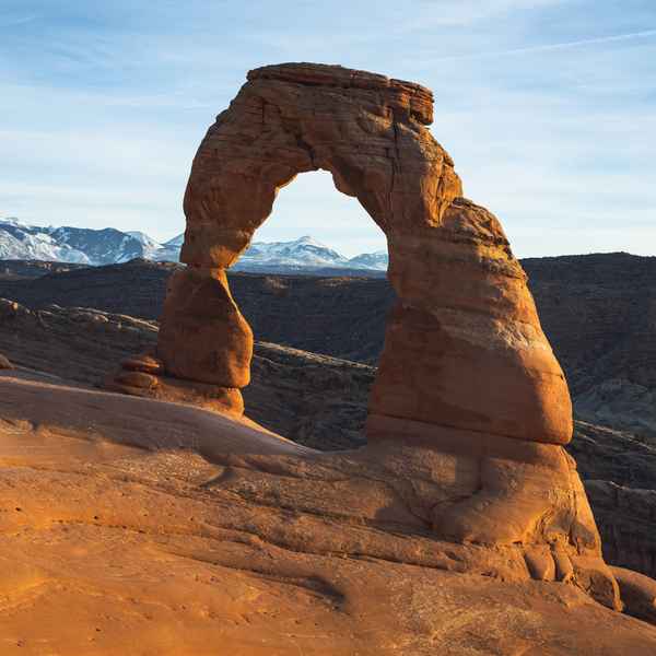 A large, sand colored arch made of rock sits on top of a ledge of the same material with snow-covered mountain peaks in the background