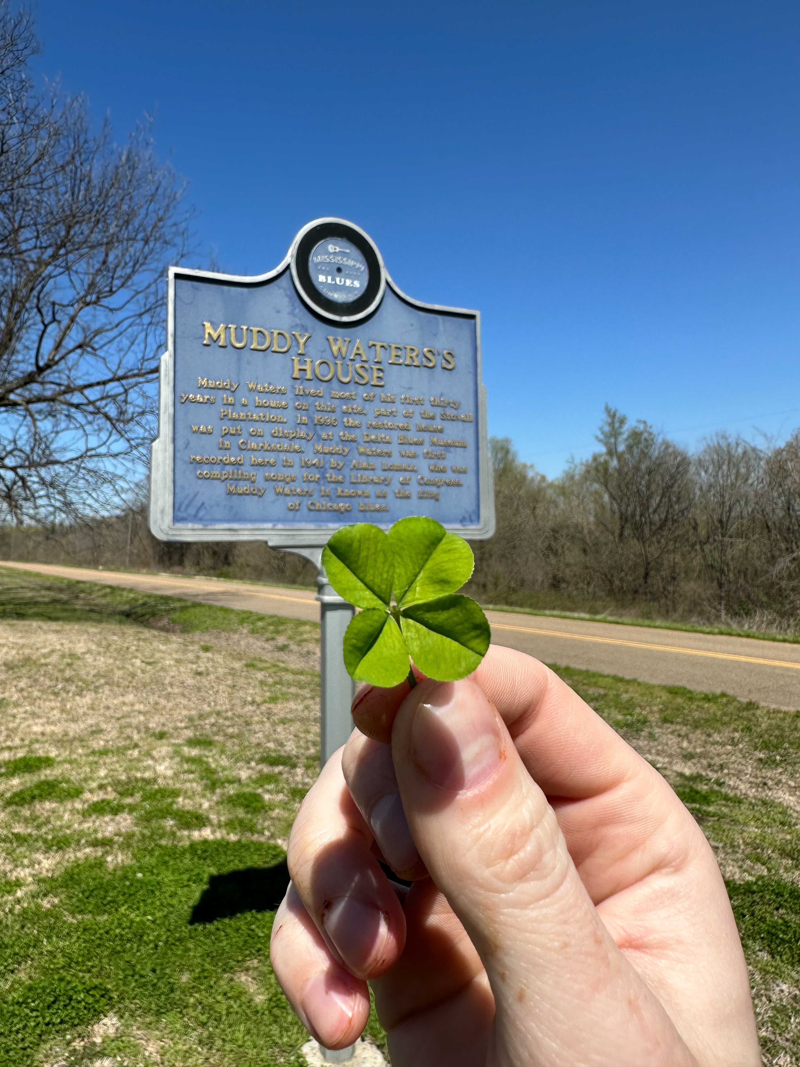 A photograph of a hand holding up a four-leaf clover in front of a historical marker that reads 'Muddy Waters's House' next to a country road