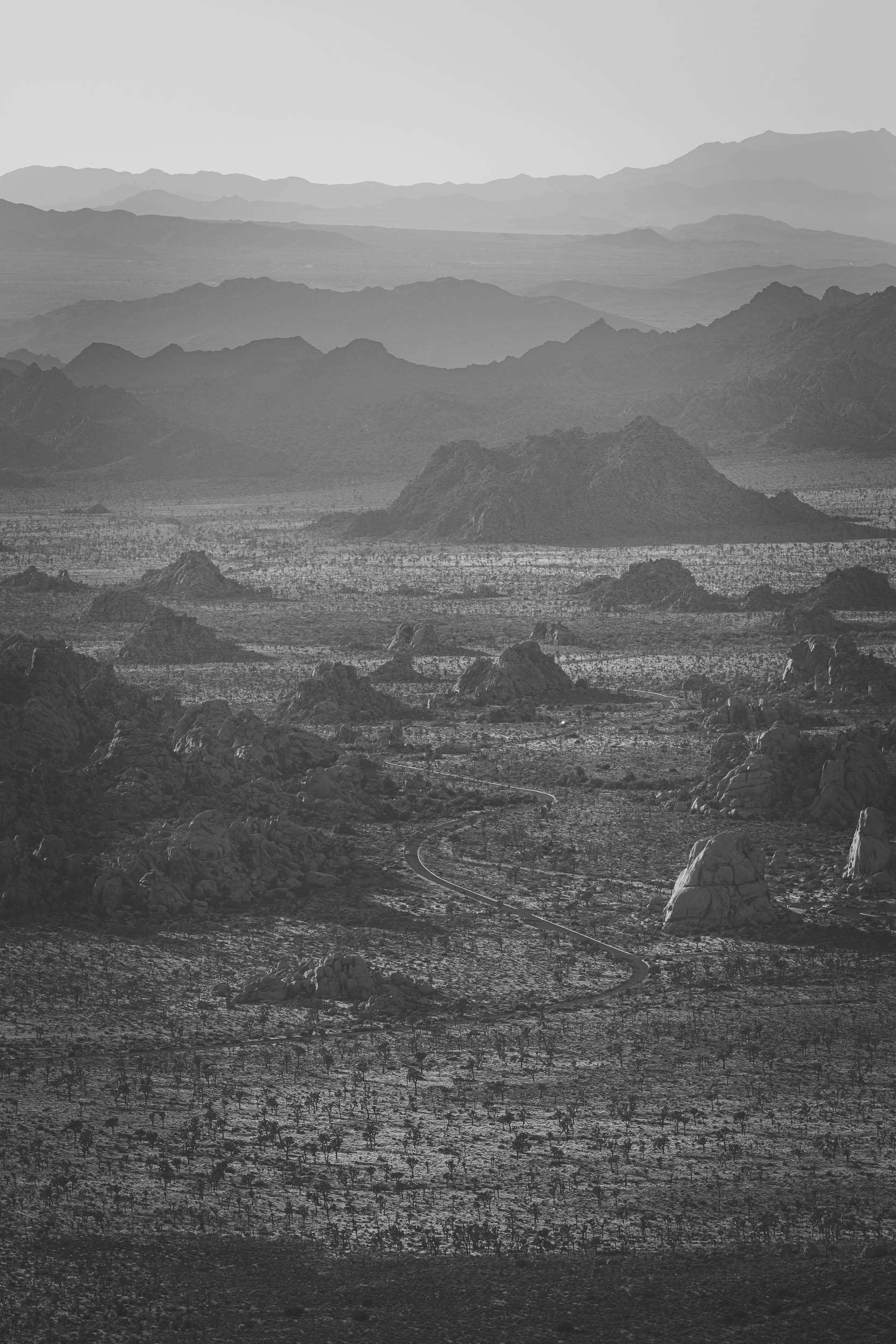 A black and white photo from a mountaintop of a road winding through rock formations on the desert floor