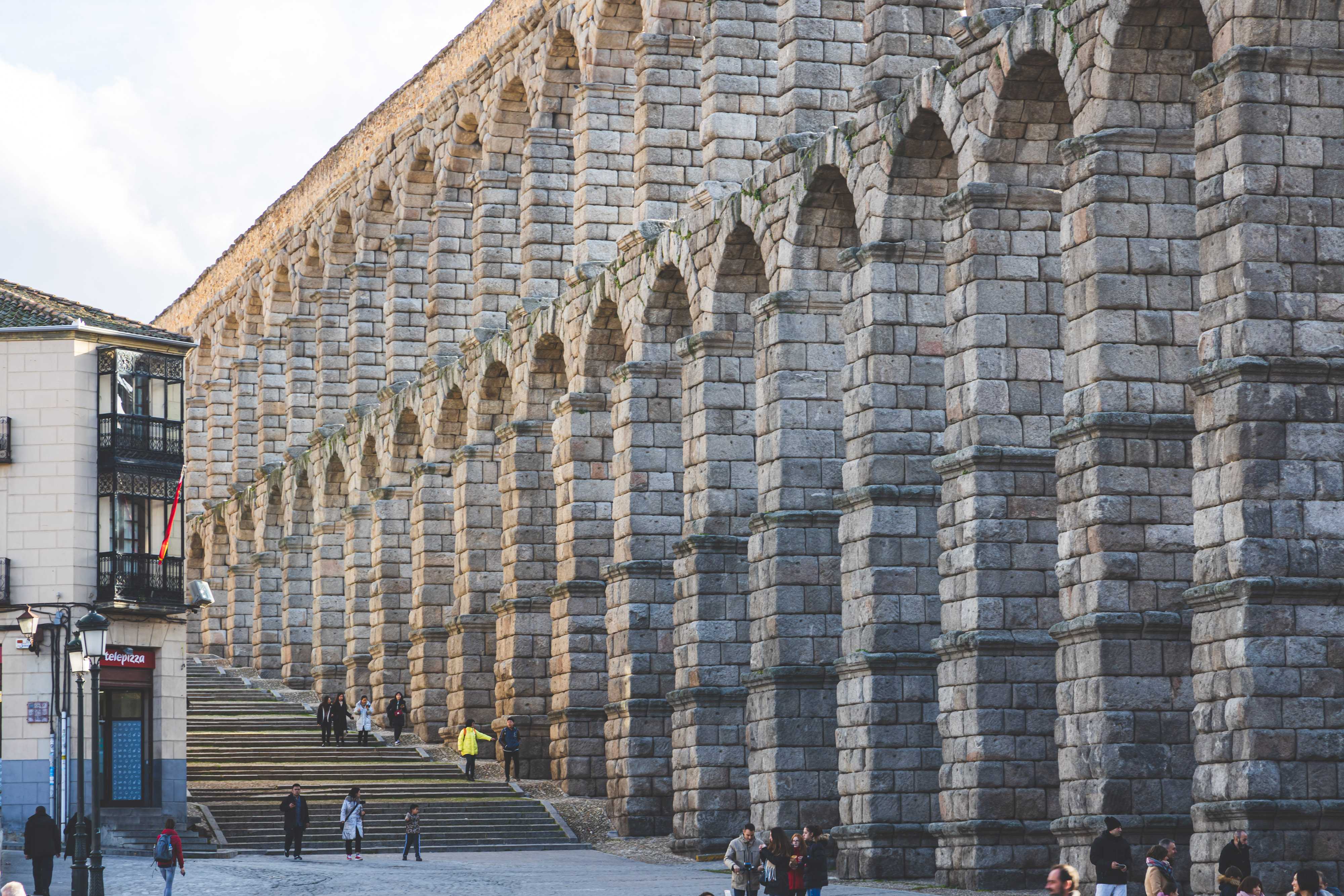 A massive Roman aqueduct bathed in golden evening light, showing its massive stone blocks that form two stacked sets of tall arches comprising its height. People can be seen walking around its base, and climing wide stairs that run parallel to it, made to look tiny by comparison.