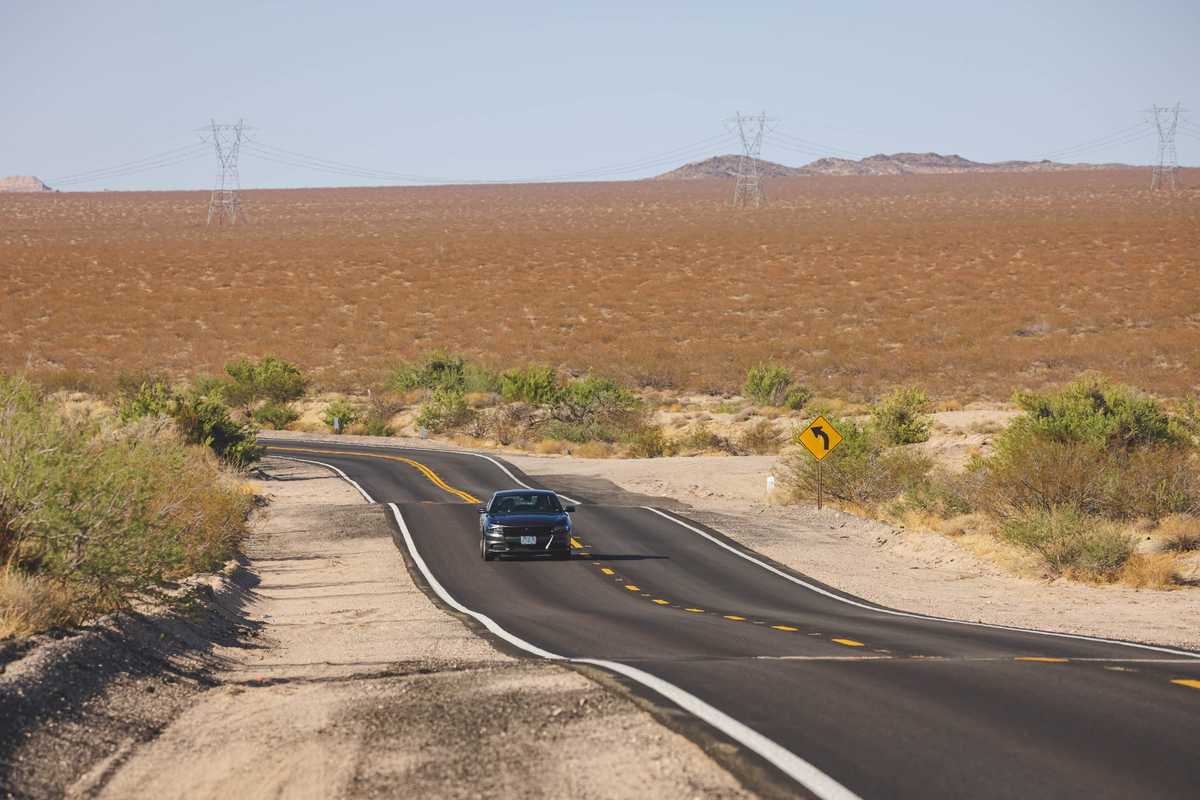 A black car drives over undulating desert roads toward the camera