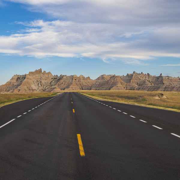 Looking down a black highway with bold yellow and white striped lines, running into the distance where it seems to meet a large, sharp, rock formation that looks like mountains on the horizon. The sky is light blue with a mass of fluffy white and light grey clouds.