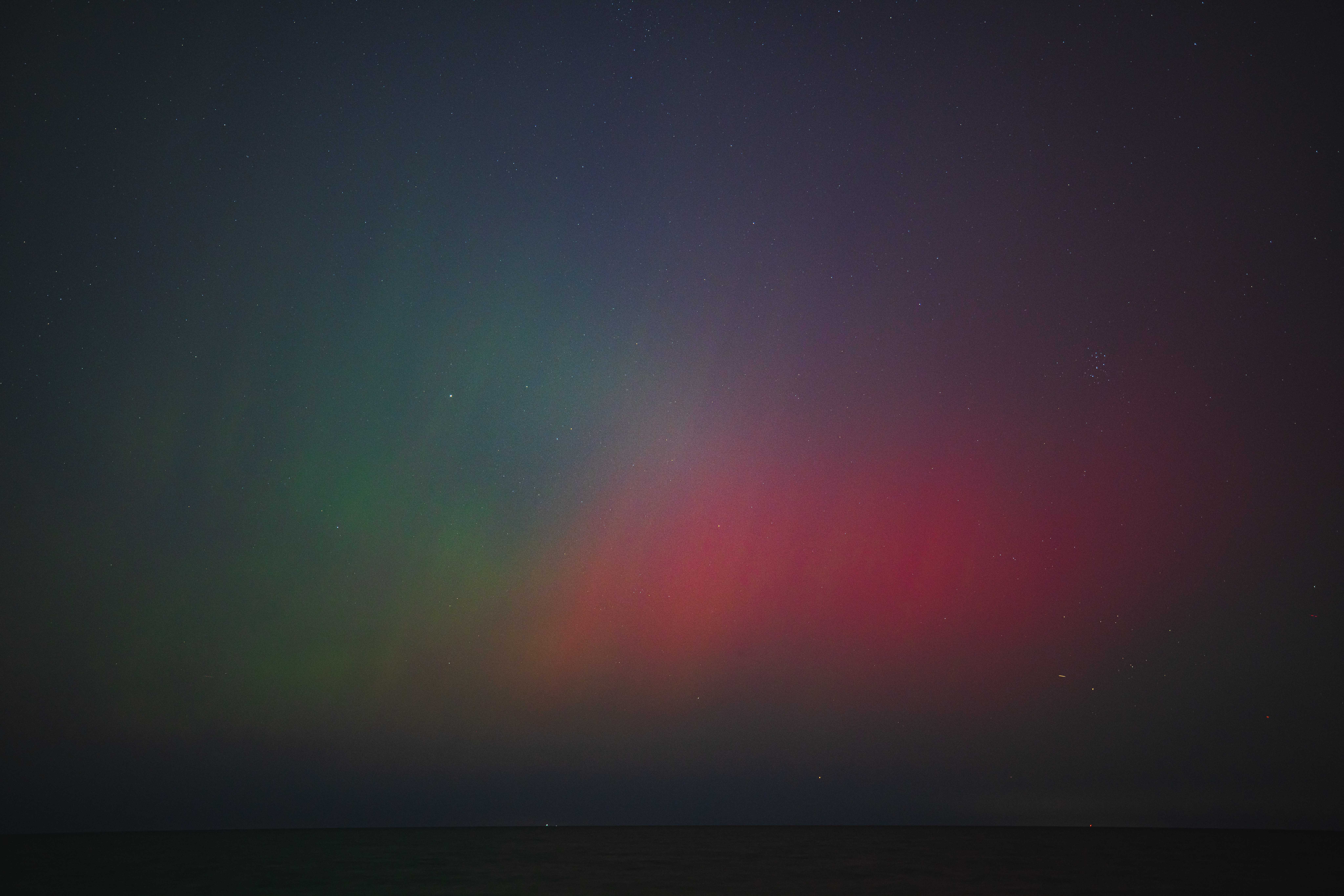 The night sky with many stars visible is glowing with green streaks on the left mixing with red streaks on the right, visible above a thin strip of Lake Michigan.