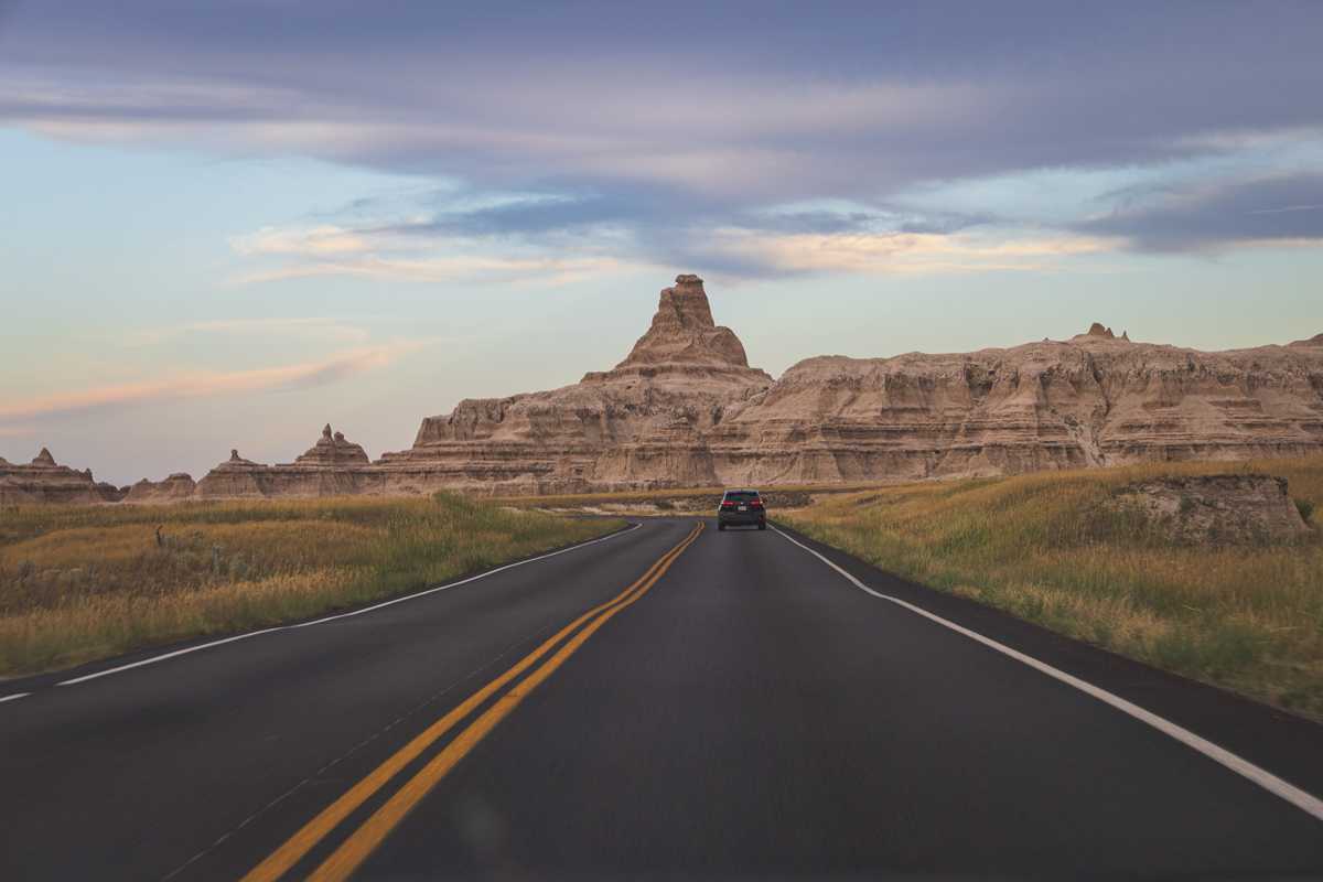 Looking down a two lane road heading toward striking rock formation featuring a narrow tower rises on the horizon