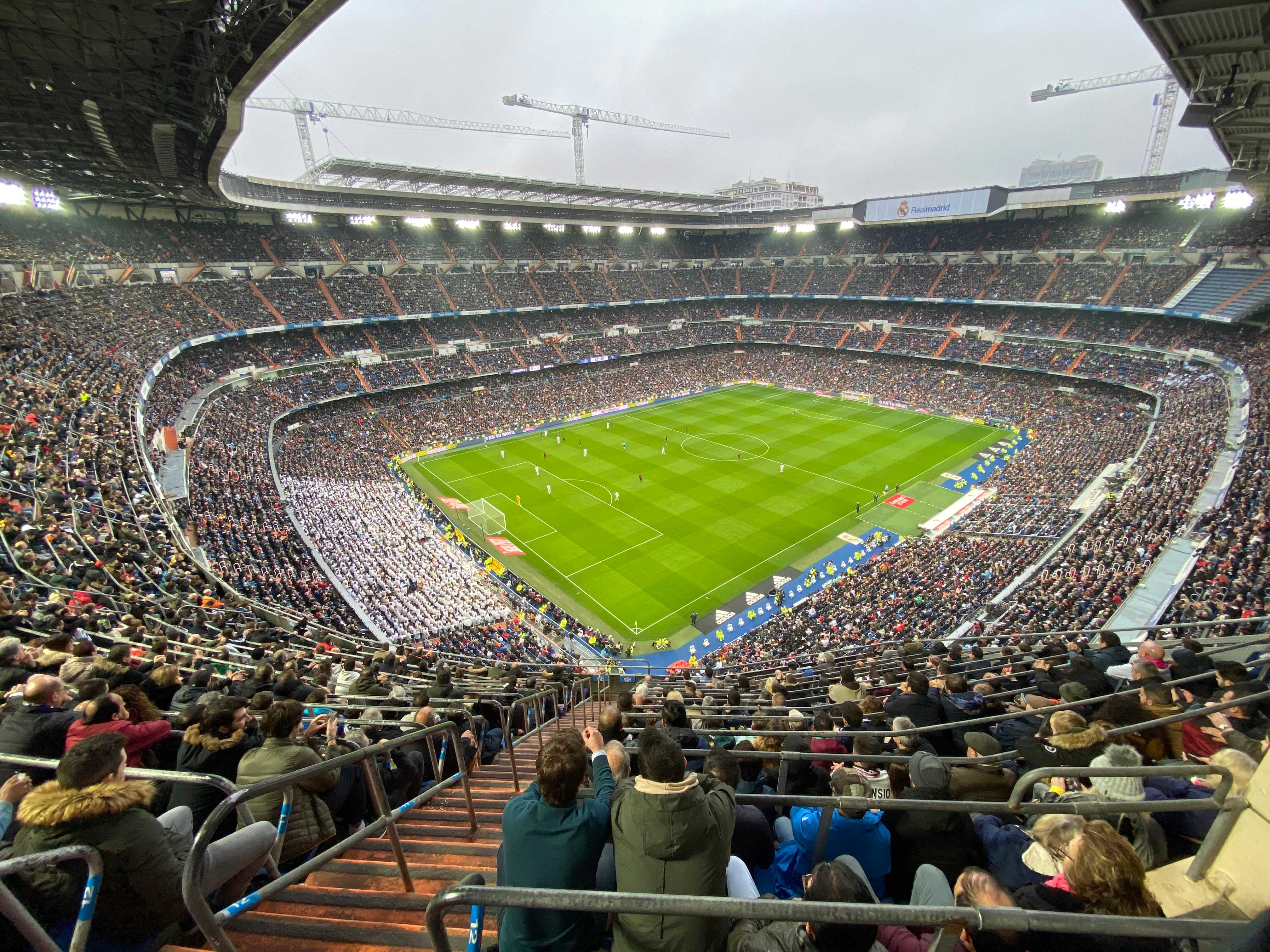 A wide angle view of Estadio Santiago Bernabéu, the stadium of the Real Madrid soccer team. The players are tiny on the green pitch, and the massive stadium is visible on all sides, filled with people.