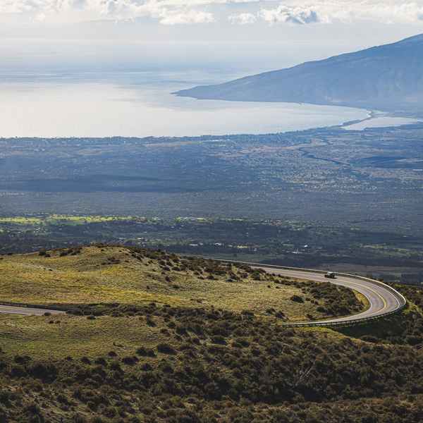 A car sweeps around a corner as a road descends a grassy, deep green mountainside, with the rest of the island of Maui and a large bay feature in the distance
