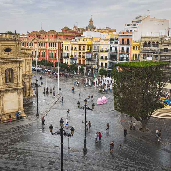 People milling around Plaza de San Francisco on a rainy day, the deep gray cement of the square shiny with rainwater. In the background, brightly colored buildings line a street.