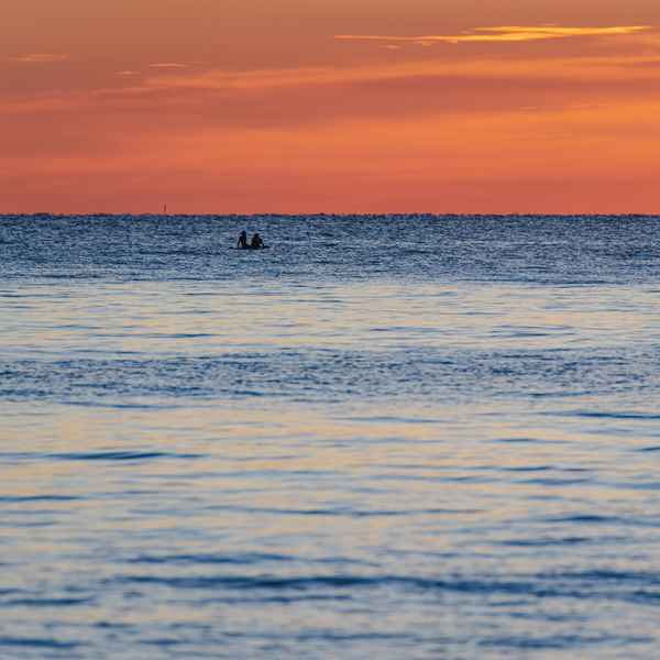 The sunrise has turned the sky orange above the deep, shining blue water of Lake Michigan, rolling gently. This cuts the photo in half horizontally between deep orange and bright blue. A pair of people are sitting on a paddleboard on the lake in the distance, watching the sunrise.