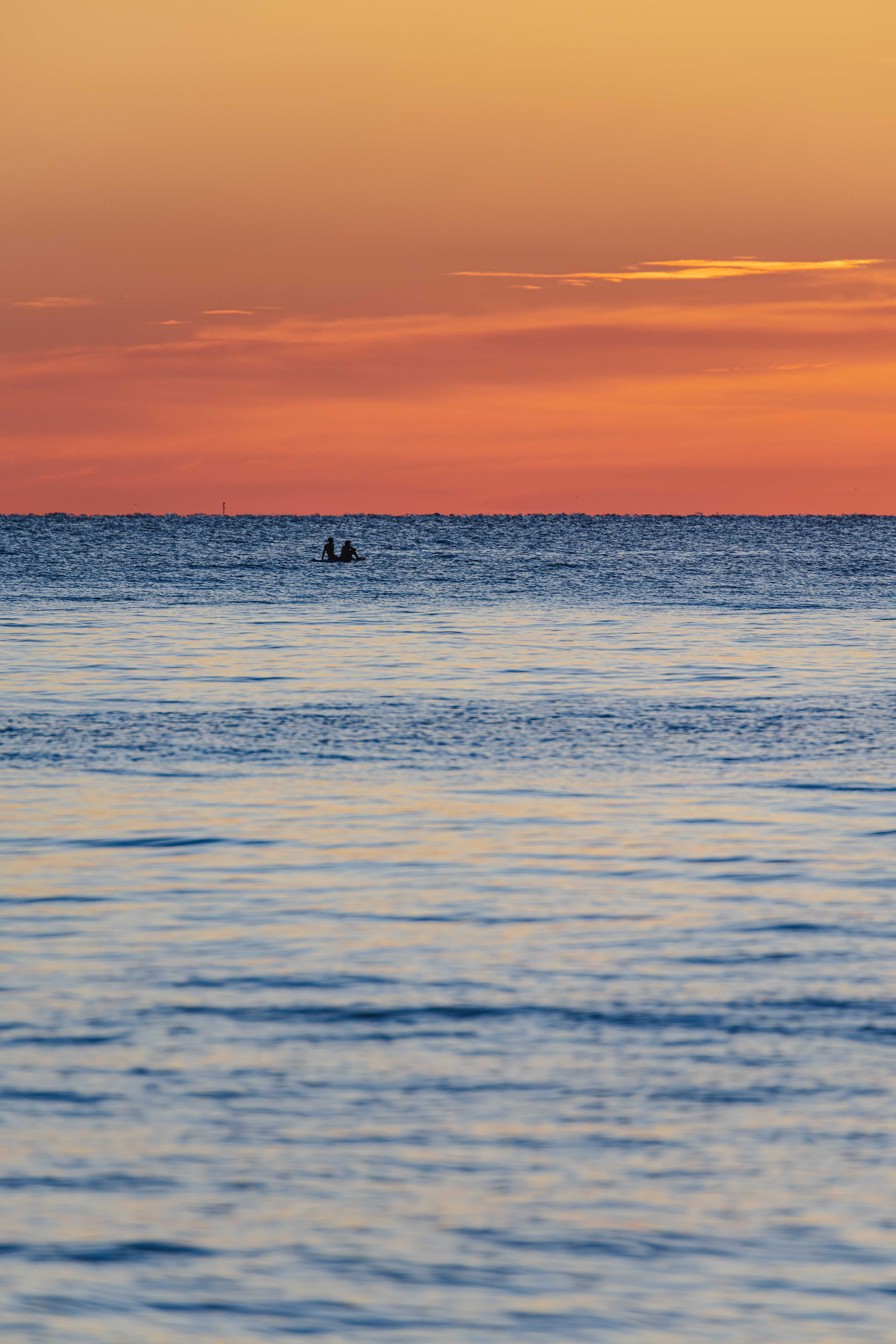 The sunrise has turned the sky orange above the deep, shining blue water of Lake Michigan, rolling gently. This cuts the photo in half horizontally between deep orange and bright blue. A pair of people are sitting on a paddleboard on the lake in the distance, watching the sunrise.