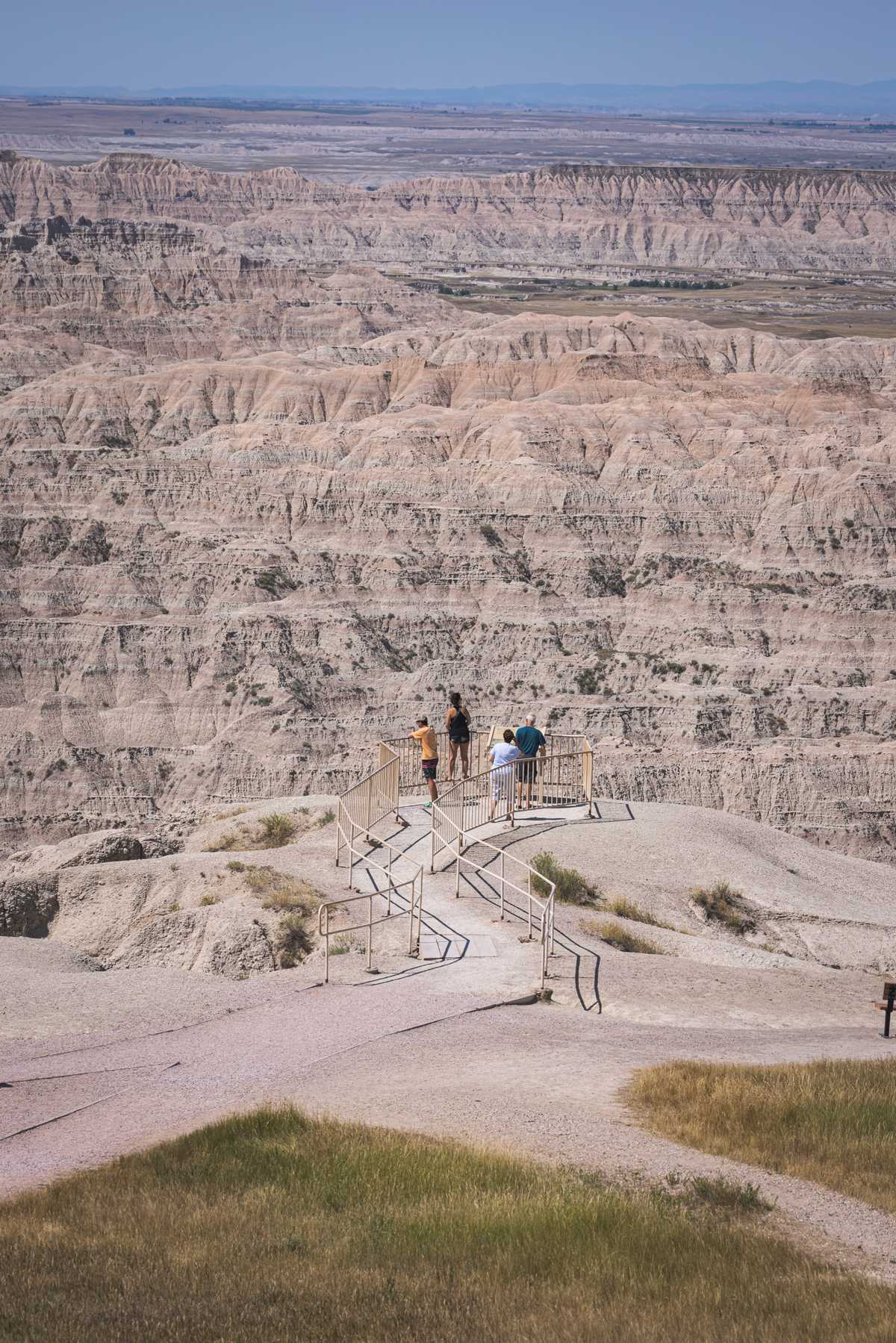 A small group of people stand in a small, fenced-off observation area on a cliff viewed from behind as miles and miles of rocky hills stretch into the distance