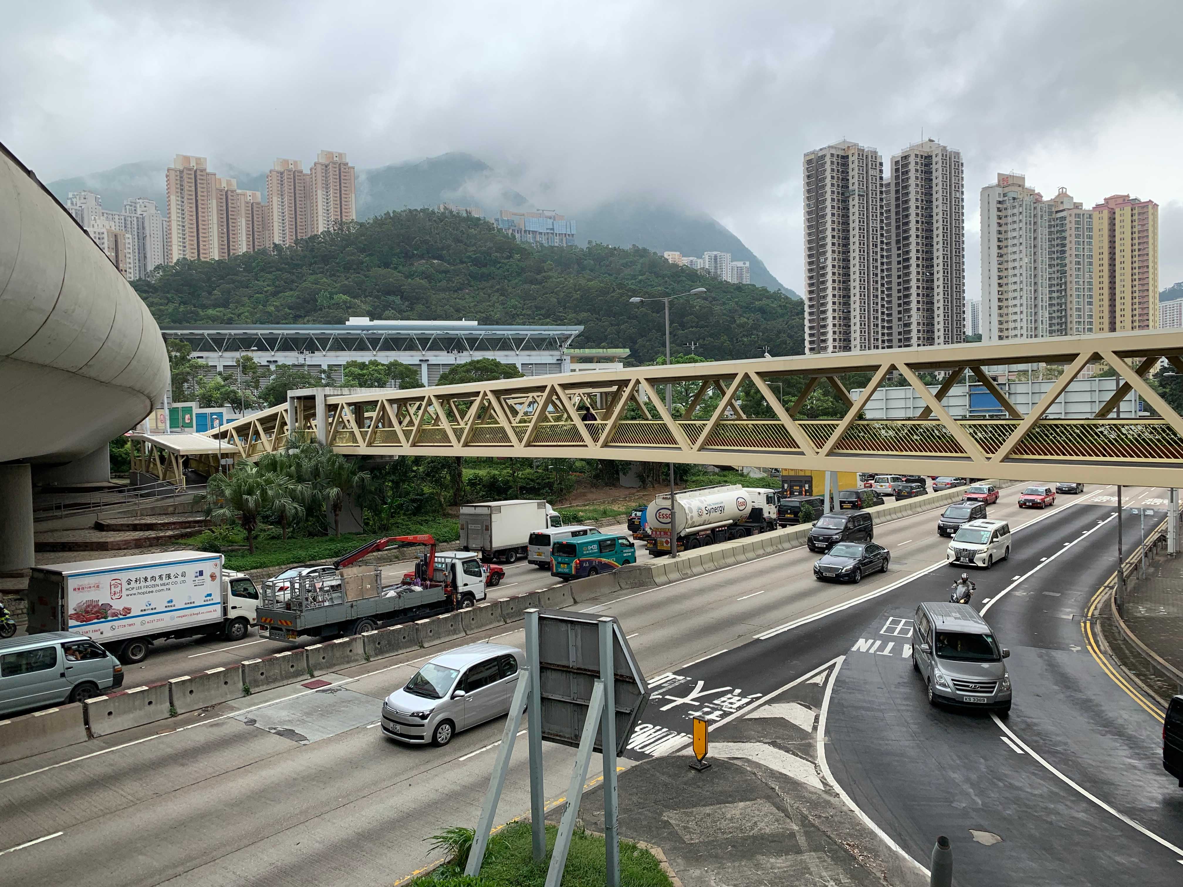 A busy scene shrouded in fog as a divided highway runs underneath the visitor, covered in cars, as a pedestrian bridge crosses the highway, spanning toward the distance, which shows some heavily forested small mountains punctured by many tall high rise buildings
