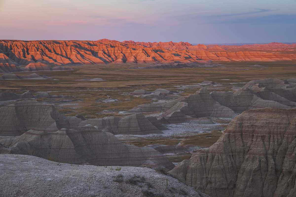 As the sun sets, a line of rocky hills is lit by warm red light, dividing a landscape between shadow, where the sun has gone below the horizon, and brightness, where its light is still peeking out