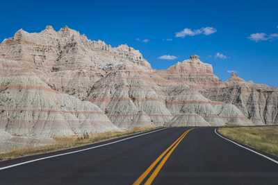 A photo of a jet black road with brightly painted double yellow lines curving around a large rock formation which has clearly visible reddish layers from different ages all underneath a bright blue sky.