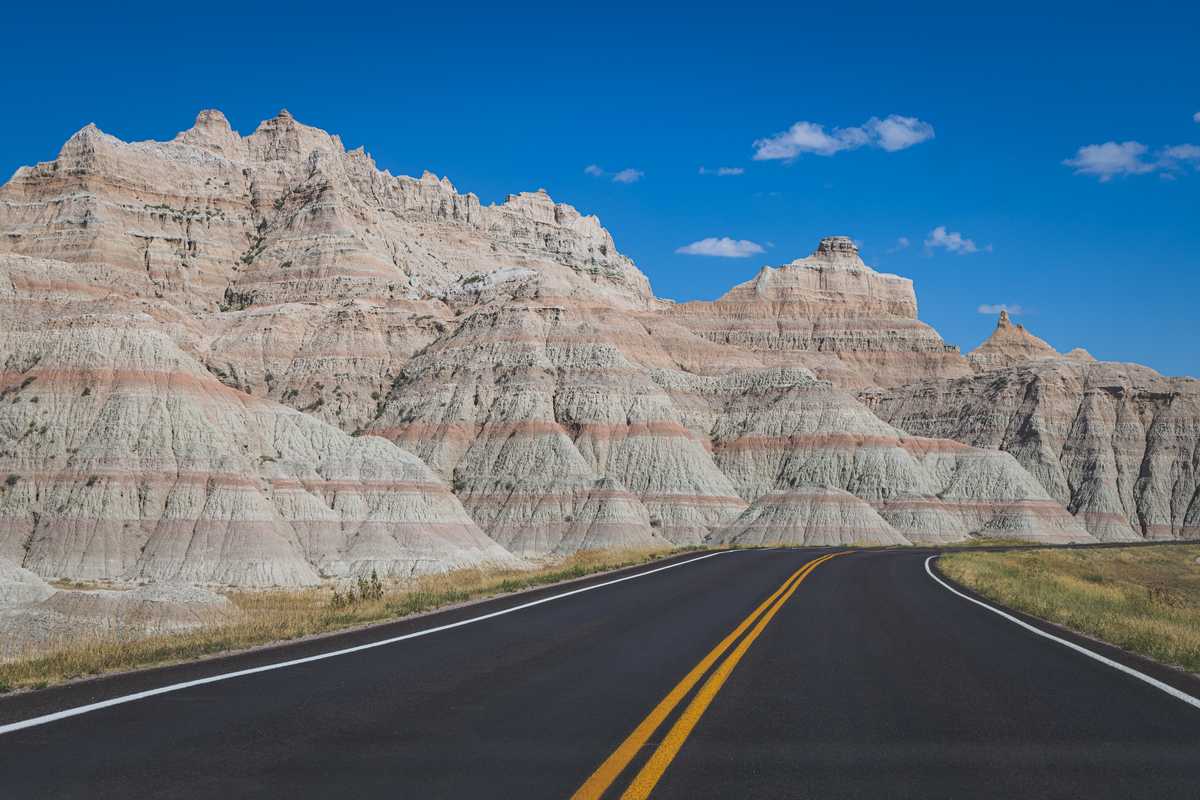 A deep black road with bright yellow lines winds to the right, around a dramatically layered formation of sedimentary rock which has bold rust-colored stripes