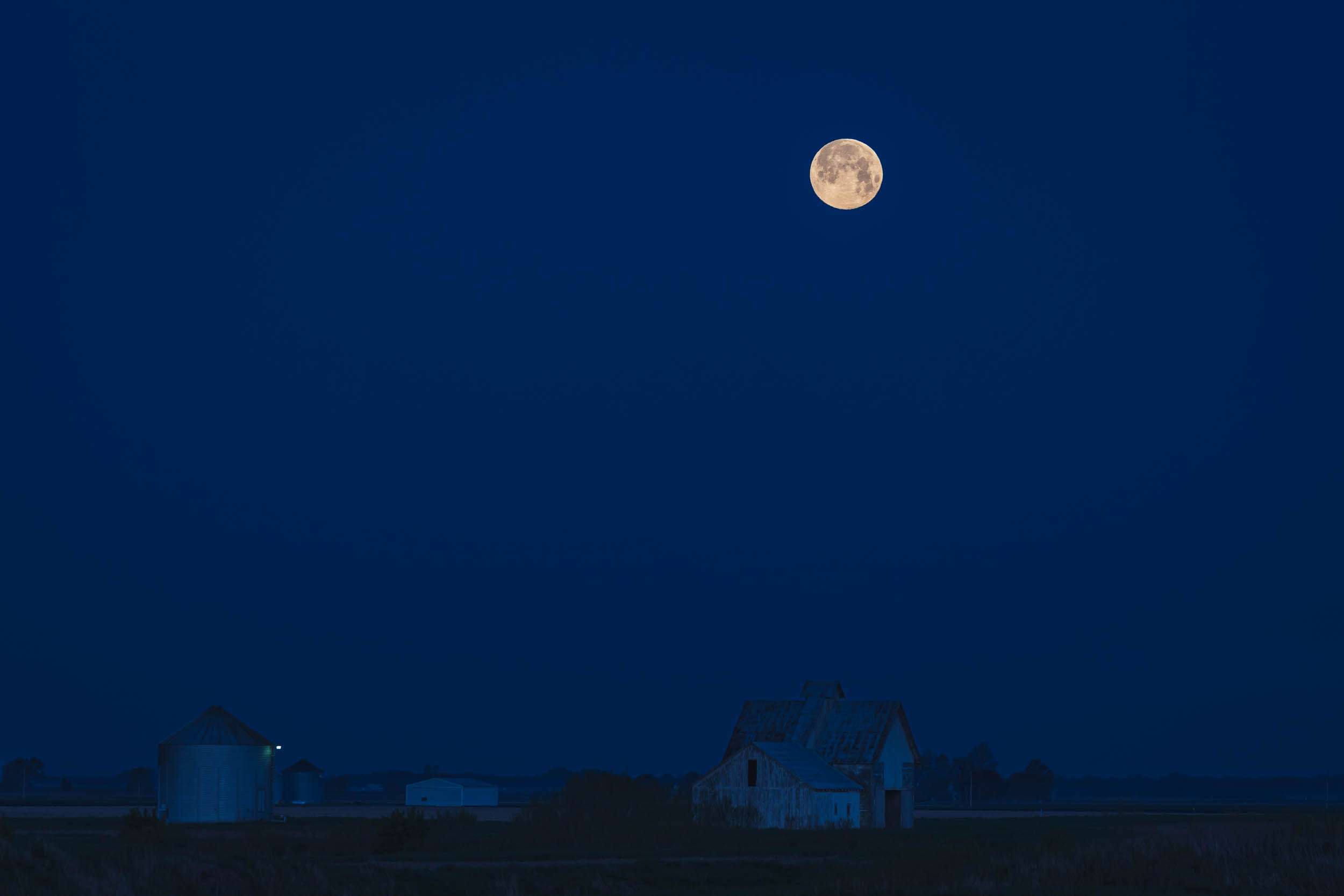 The moon shining above some farm buildings in a deep blue glow just before sunrise