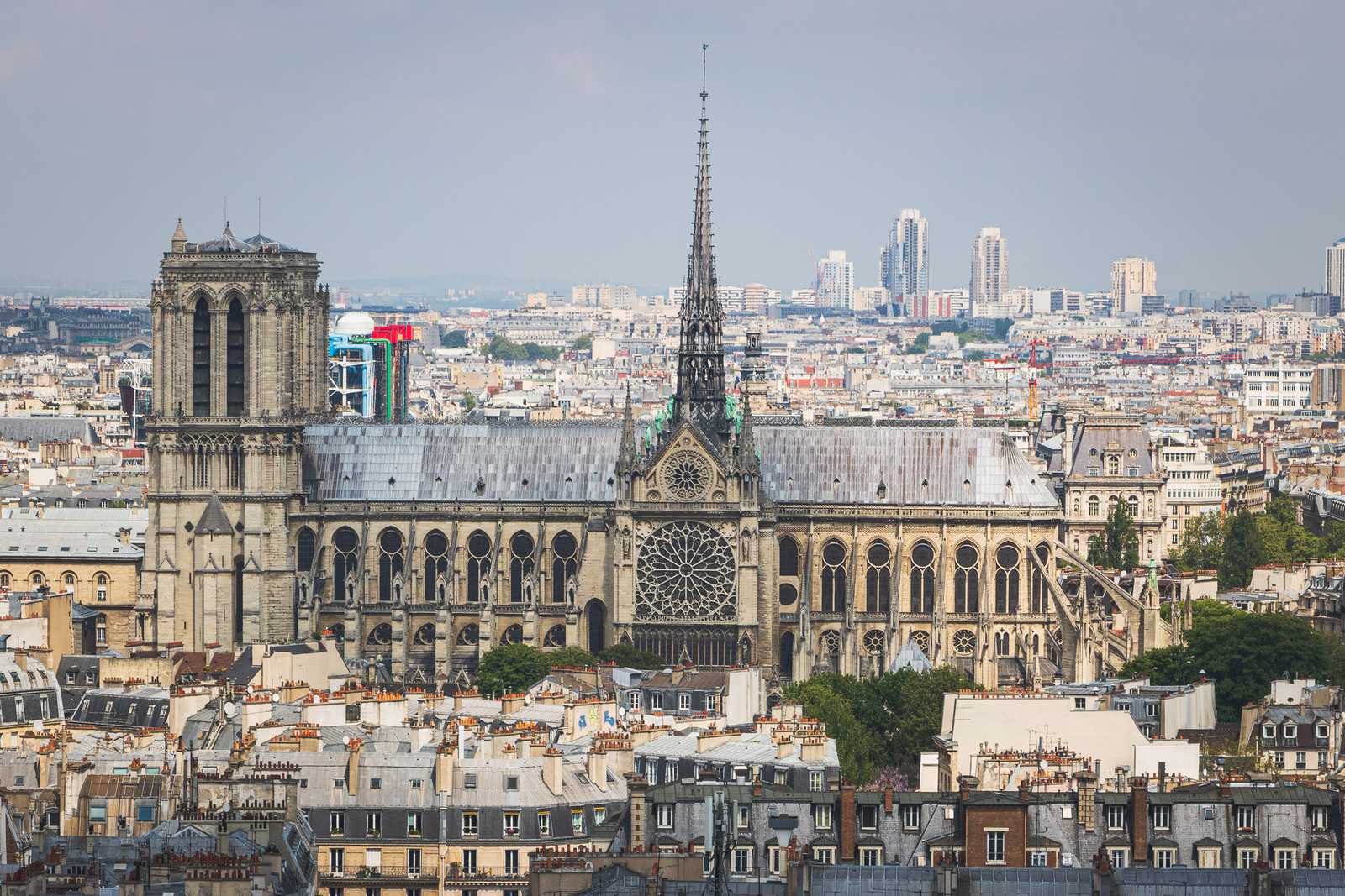 A view from slightly above the iconic towers and spire of Notre Dame, a large gothic cathedral towering above and surrounded by the smaller buildings of Central Paris