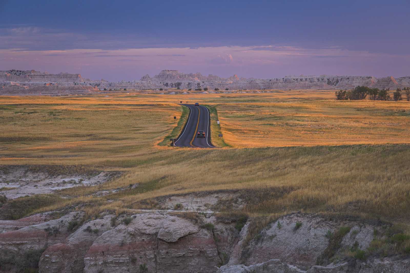 The main road in the park takes a turn, giving the viewer a sightline directly down the road as a single car drives into the park at sundown. The grasslands on either side of the road are lit with diffuse light, and the sky behind above rocky hills is a subtle purple-blue