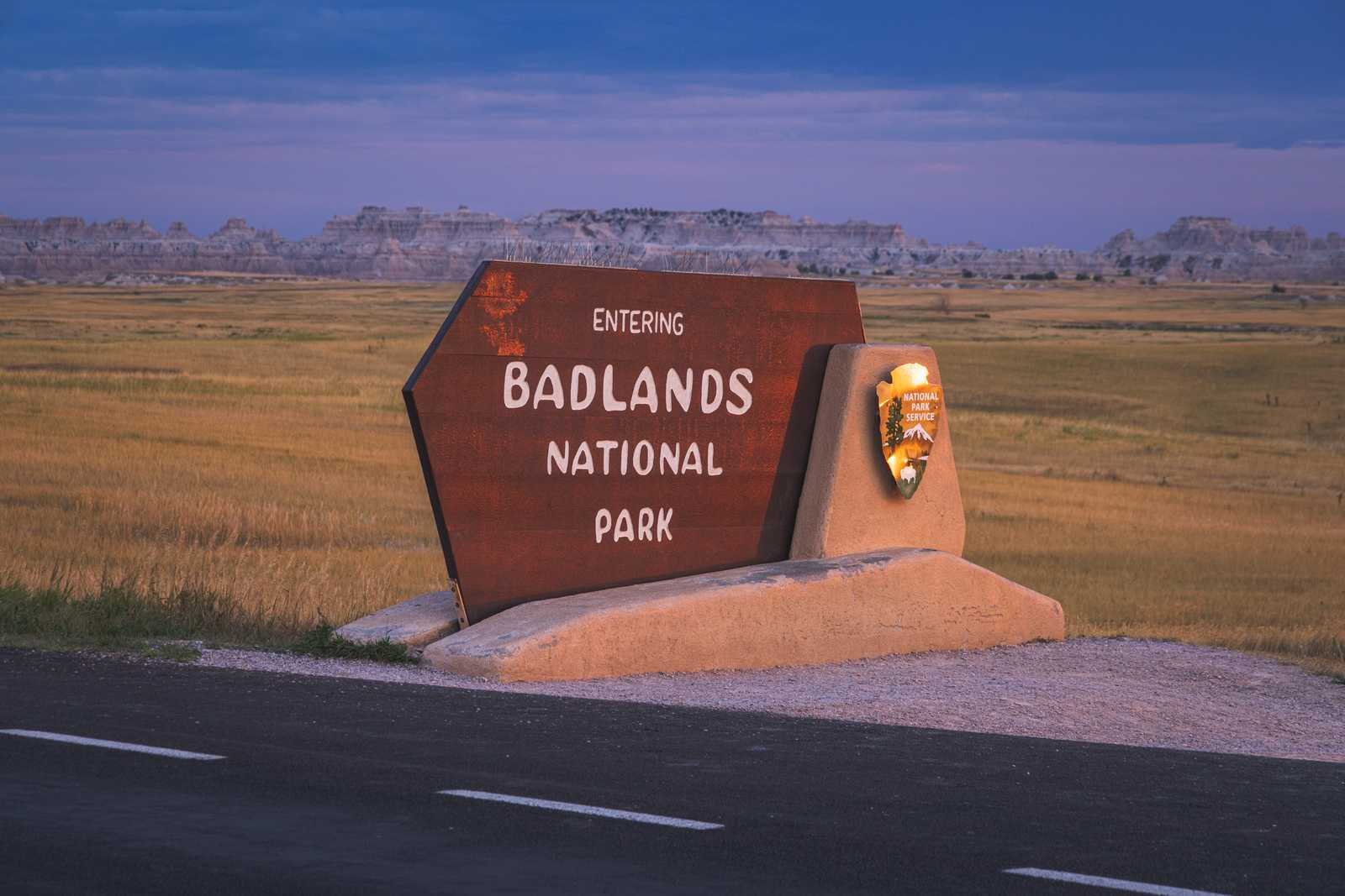 The Badlands National Park entrance sign in the characteristic rounded font of National Parks Service signs as light falls and the sky turns a purplish blue in the background