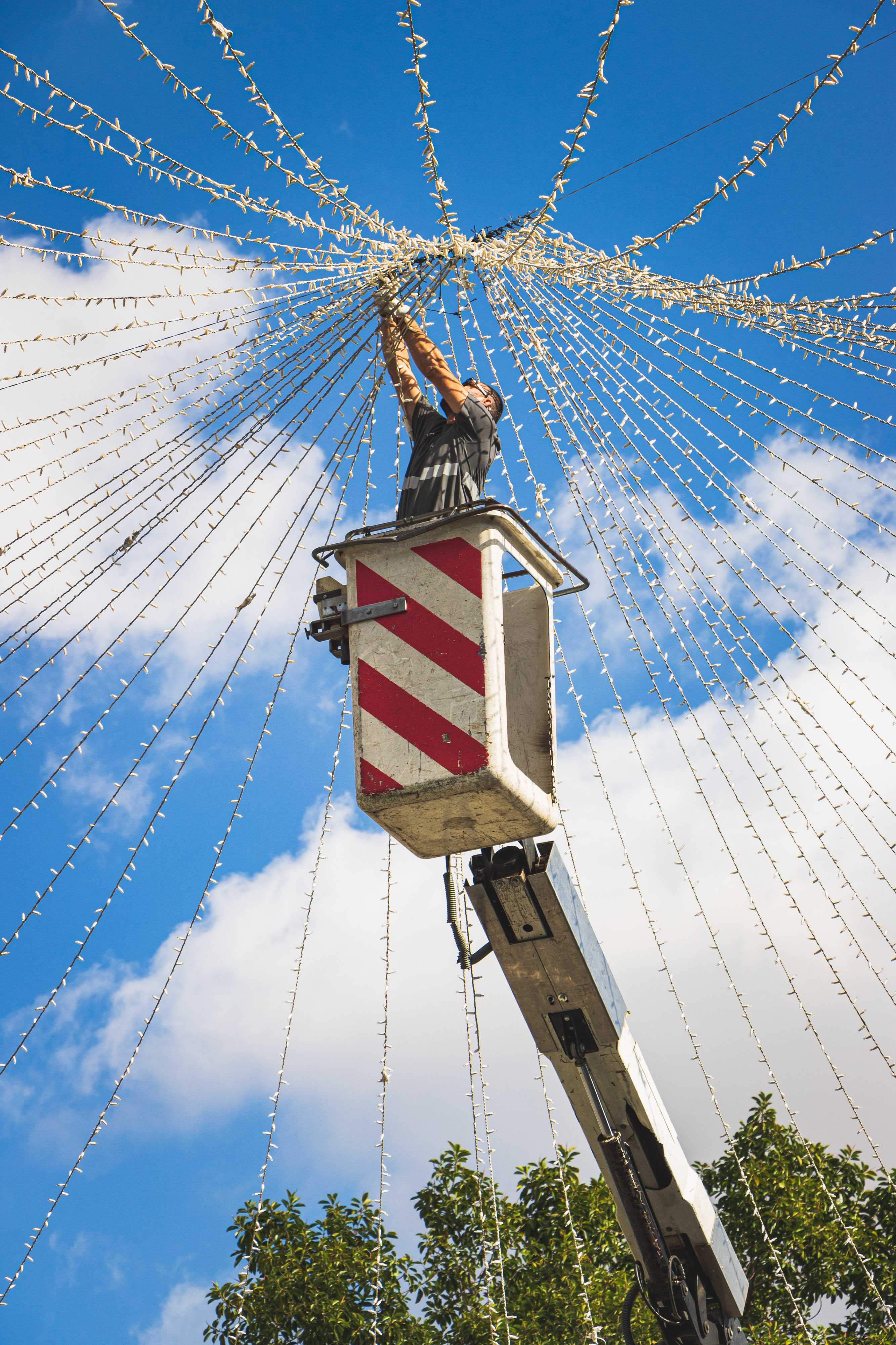 A man in a bucket lift hangs lights as they cascade down around him in a cone shape of different strings of tiny lights