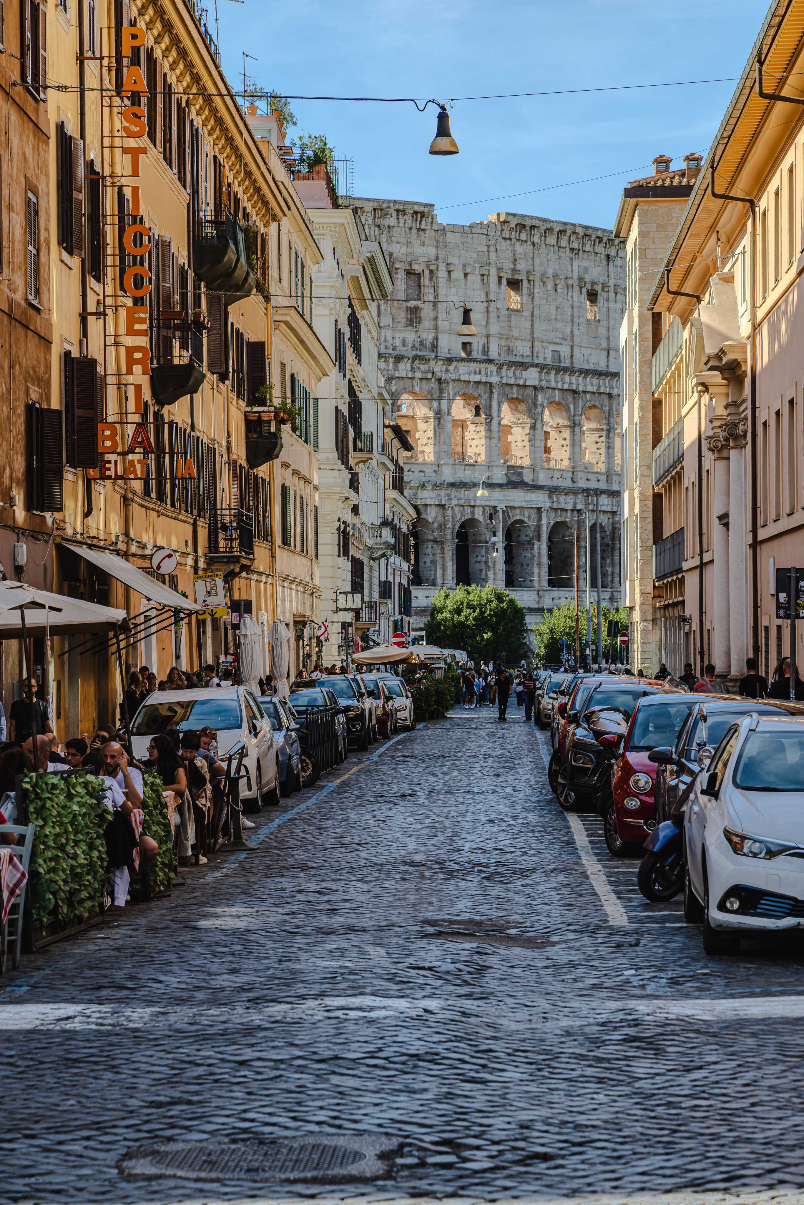 A street lined with three or four story stucco buildings leads the viewer to the Colosseum, a section visible with white ancient stone with layers arches and colums, the morning light shining through brightly from behind.