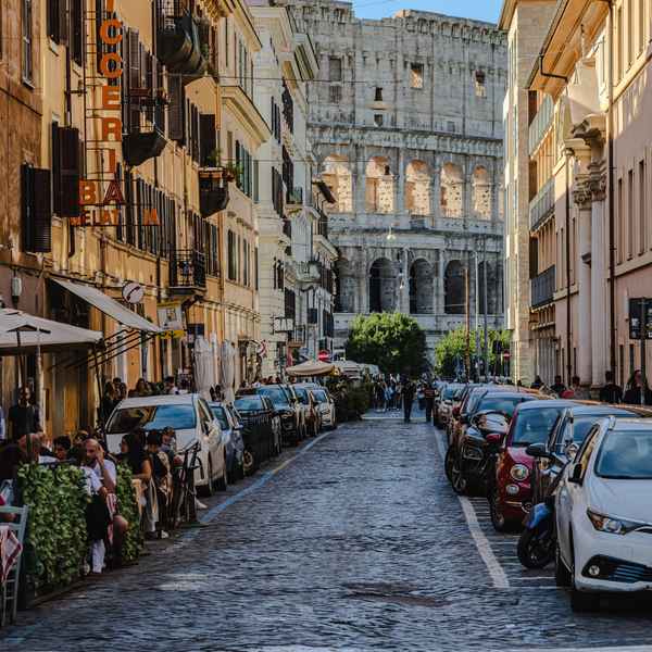 A street lined with three or four story stucco buildings leads the viewer to the Colosseum, a section visible with white ancient stone with layers arches and colums, the morning light shining through brightly from behind.