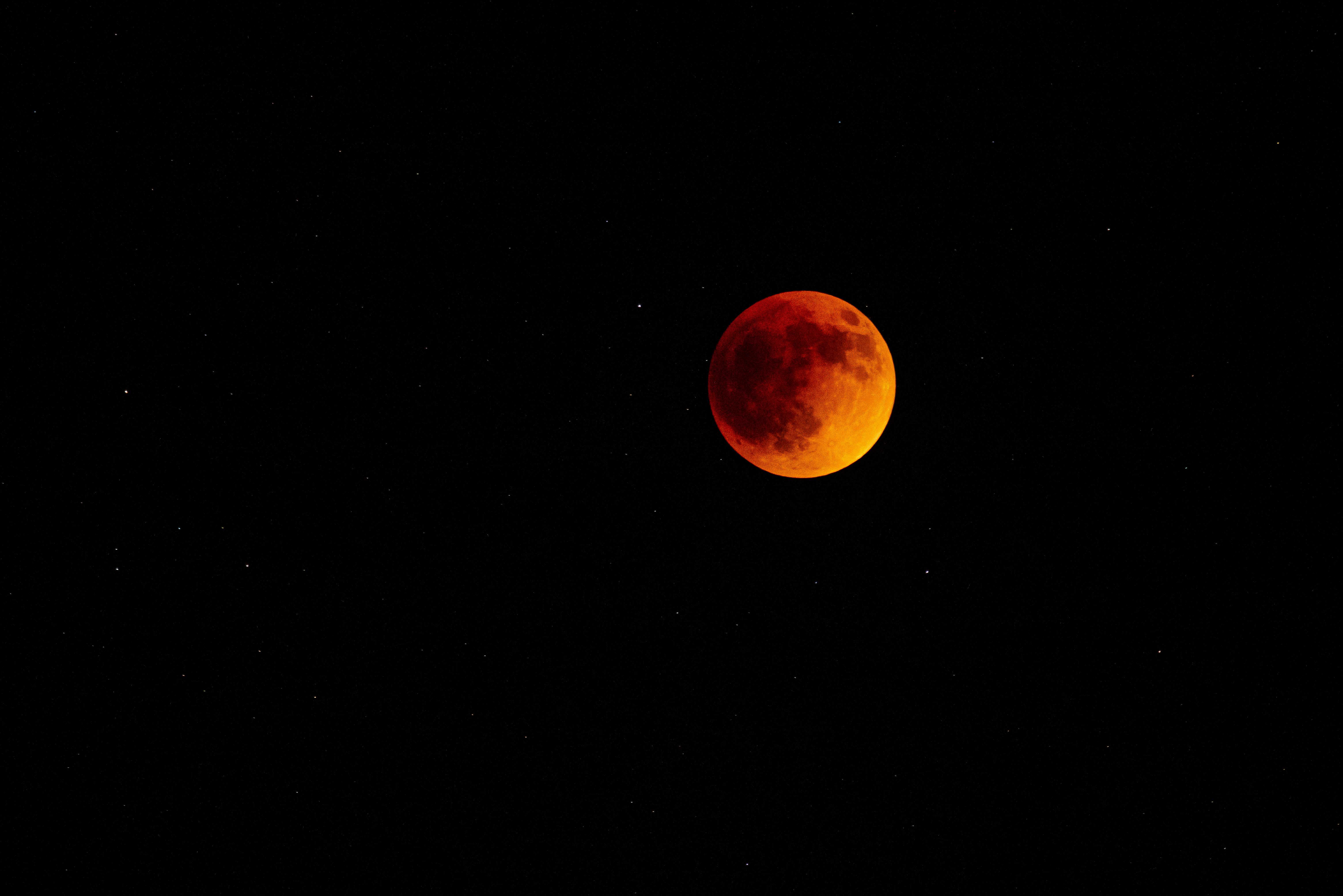 The full moon at a total eclipse shines a dull orange in the sky surrounded by stars.