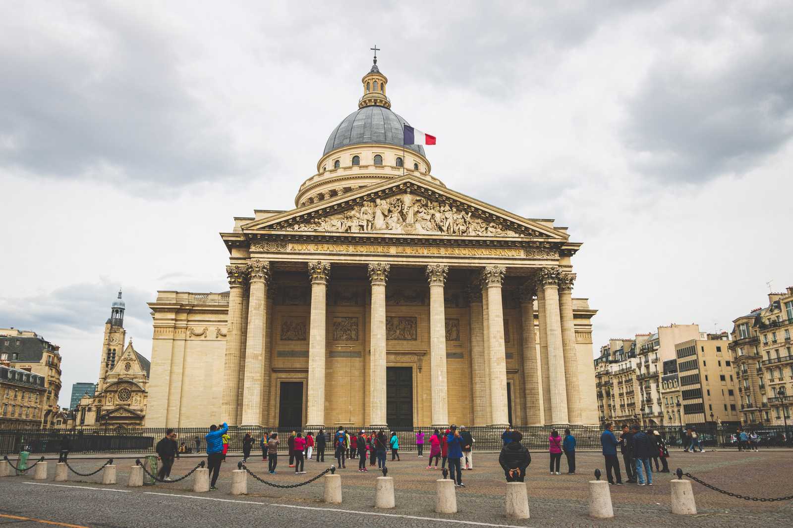 Looking up at the massive edifice of the Panthéon, Romanesque columns towering up from the plaza that surrounds it, below a massive dome behind prominent French flag