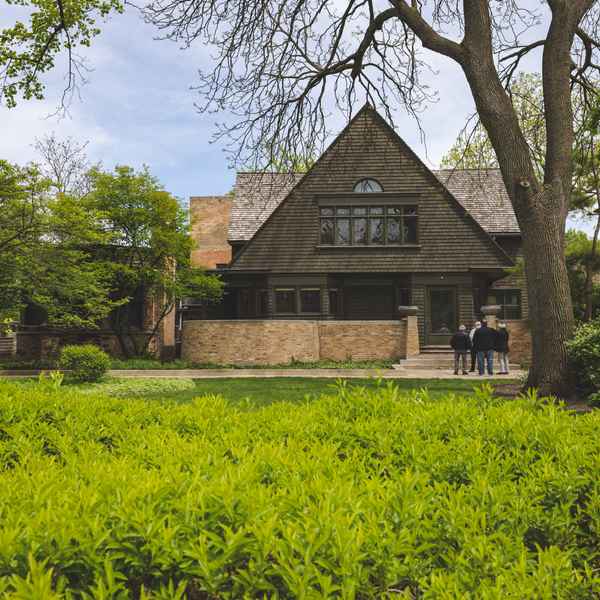 The deep brown stone facade of a neatly angular house is contrasted by the bright green of its yard, while a small group of people stand and examine its features.