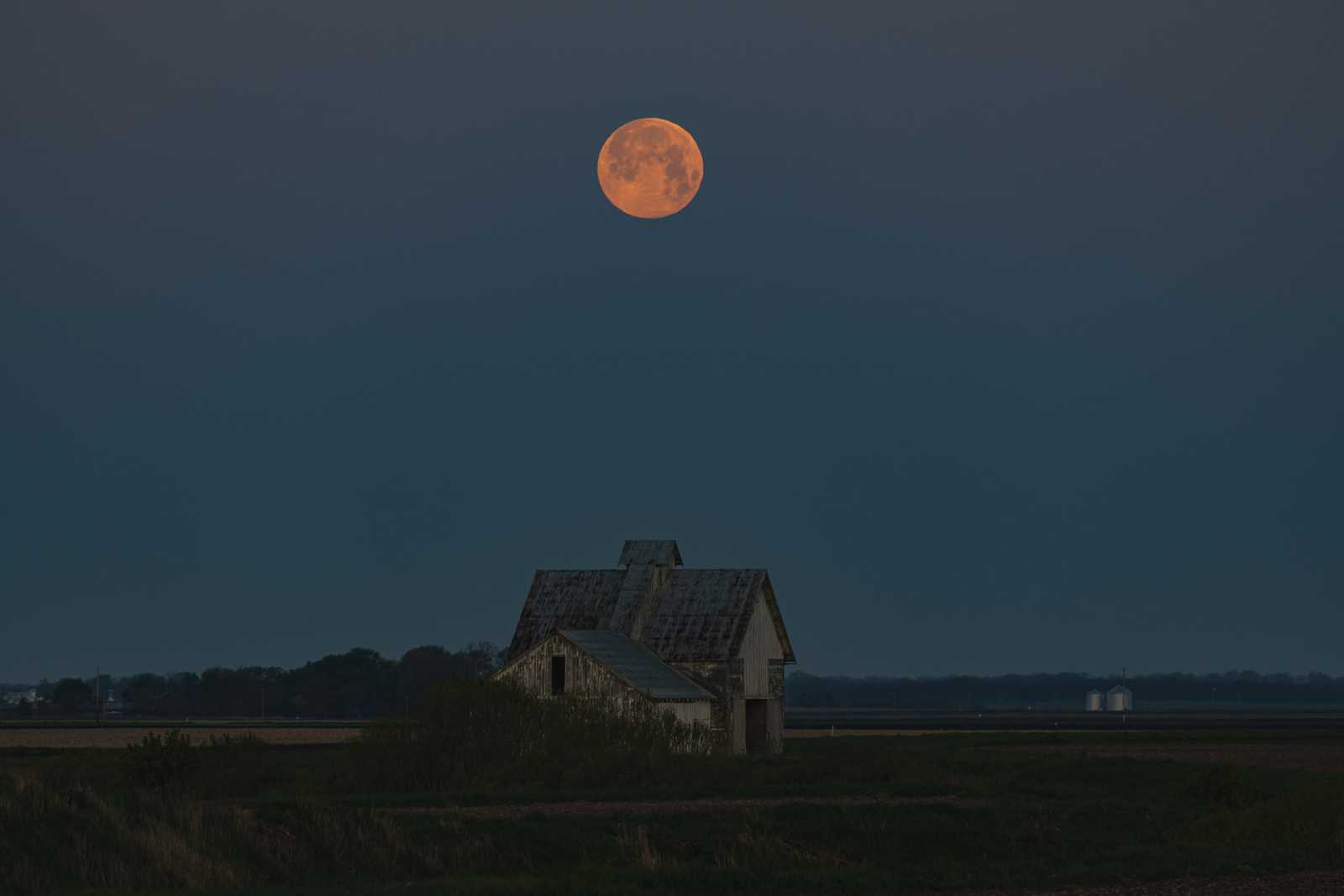 Another photo: the moon sets as dawn brightens the sky above some farm buildings in a field