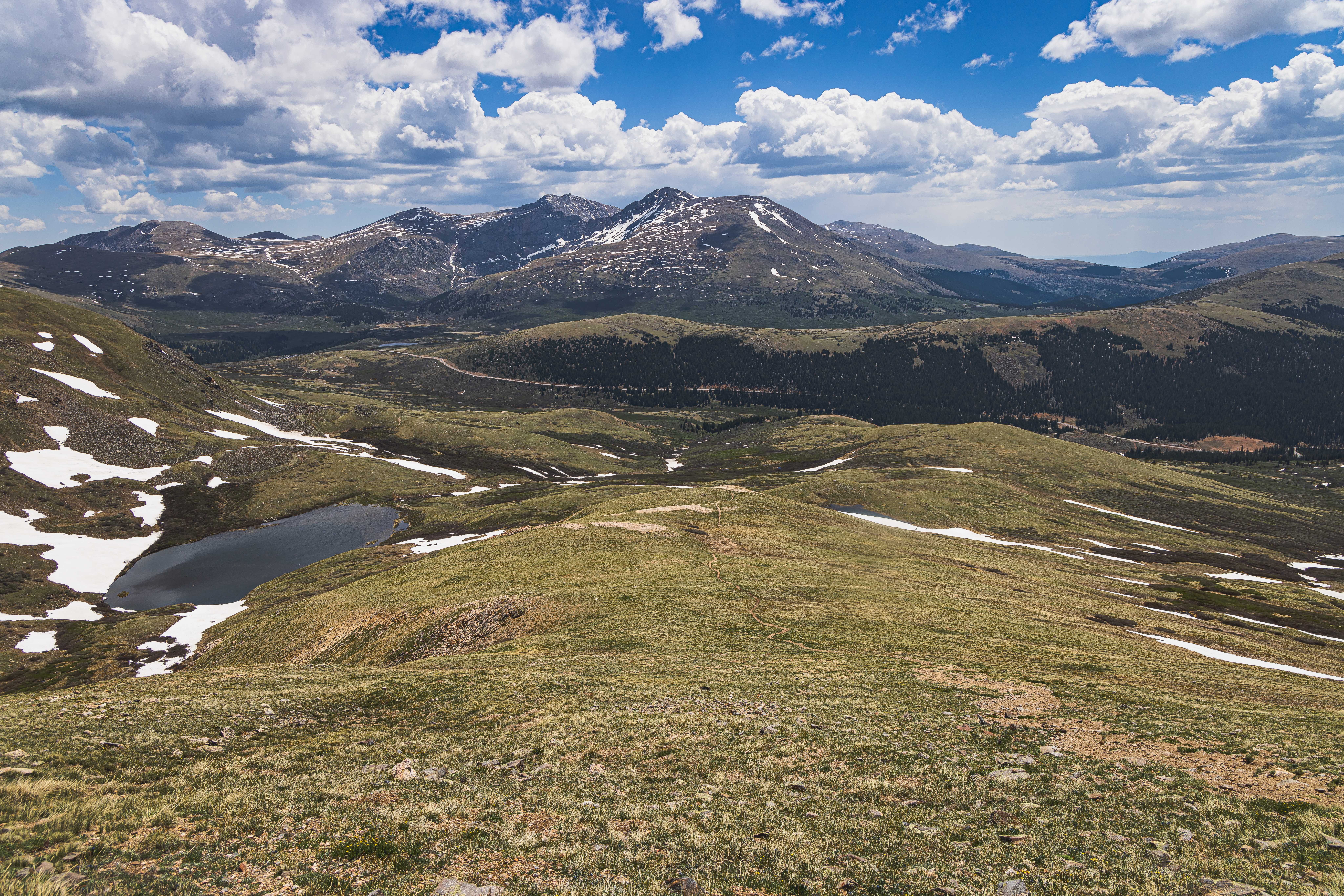 A small dirt path winds down a mountainside into a rolling green valley, an oval Alpine lake to the left, and directly ahead across the valley numerous rocky mountain peaks can be seen. Everything is covered in scattered small patches of snow.